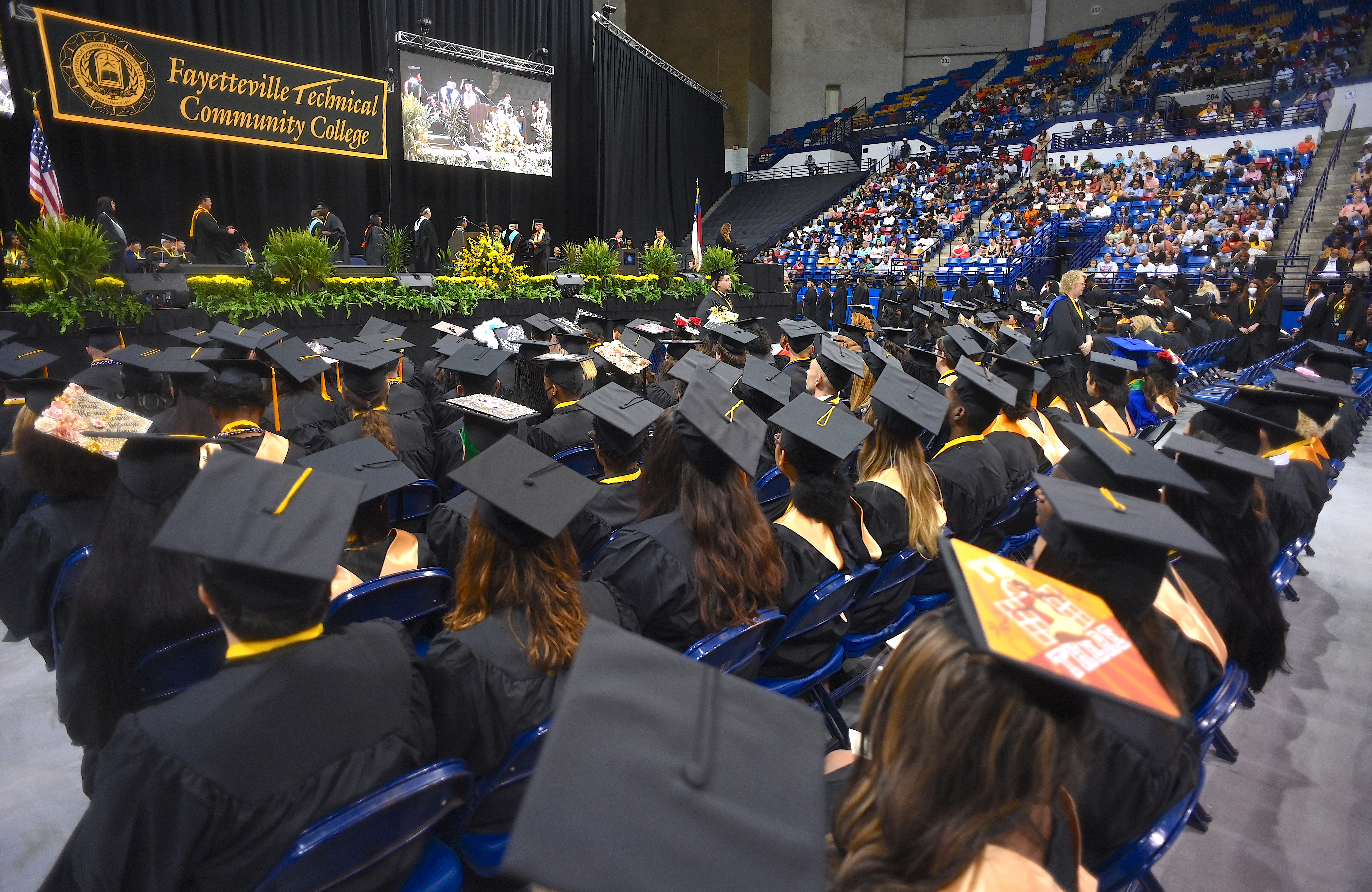A wide photo of seated graduates from behind. The stage, college banner and projection screen are visible in the background.