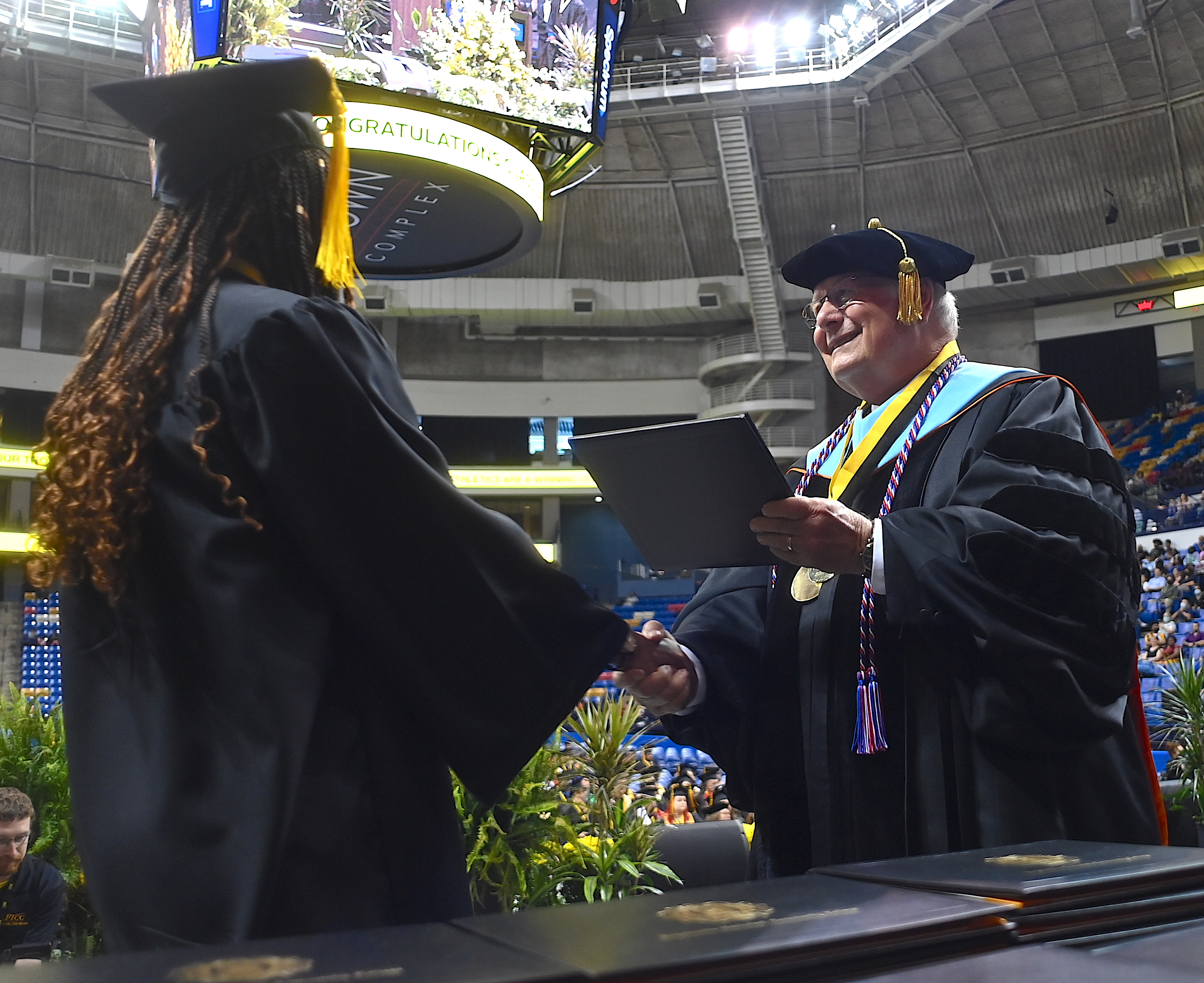 A graduate, photographed from behind, accepts her degree portfolio from Dr. Keen. 