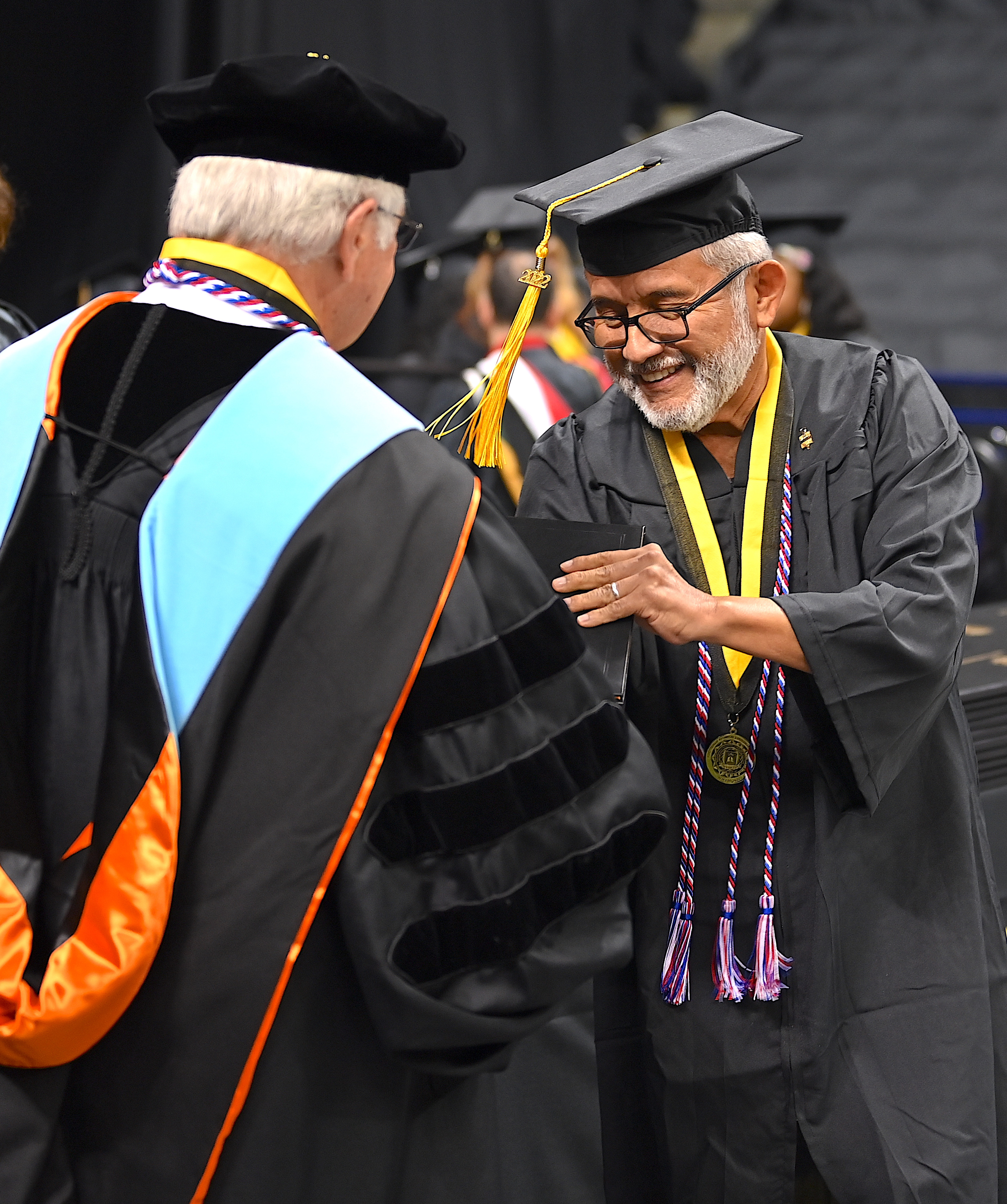 A graduate with glasses, a white beard, and a red, white and blue cord accepts his degree portfolio from Dr. Keen.