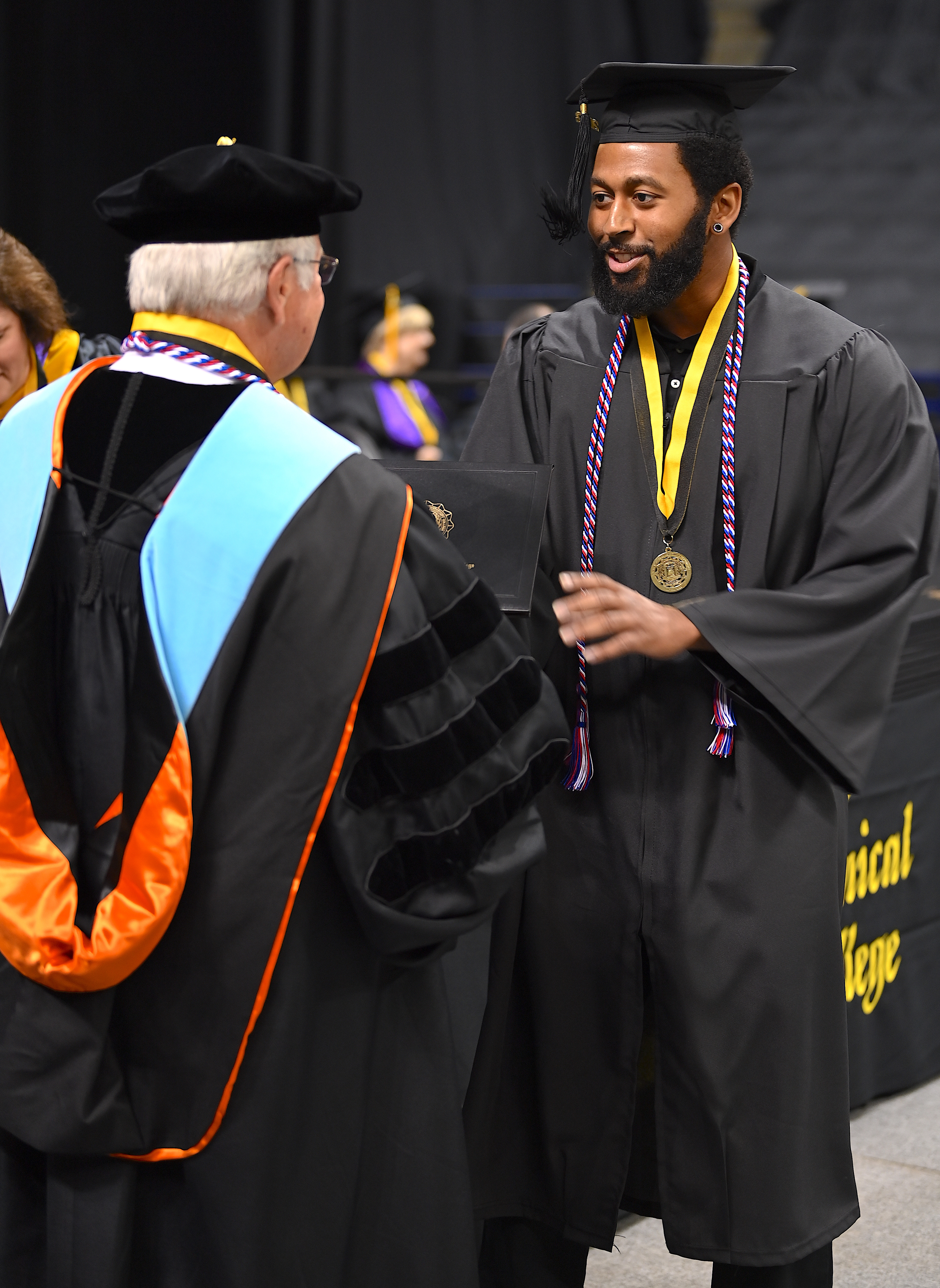 A tall graduate accepts his degree portfolio from Dr. Keen. He is wearing a red, white and blue cord and a medal on a black and gold ribbon around his neck. 