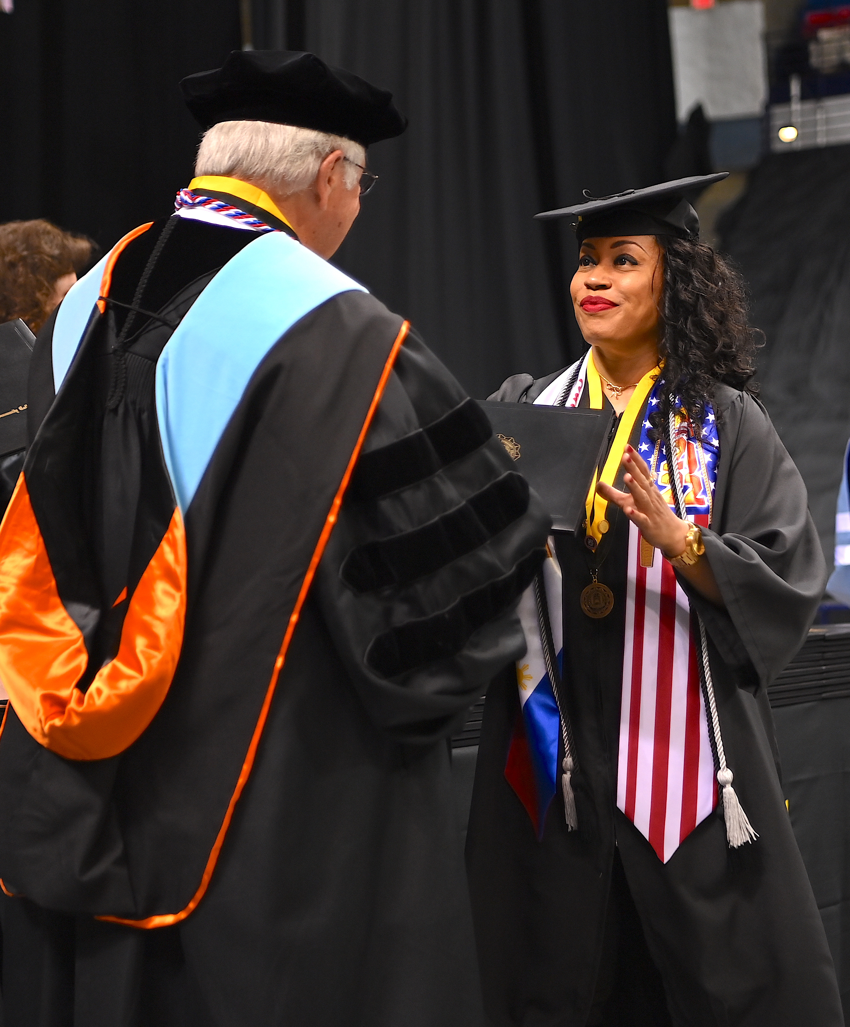 A graduate accepts her degree portfolio from Dr. Keen. She is wearing a stole with red and white stripes and stars on a blue block, symbolizing the American flag.
