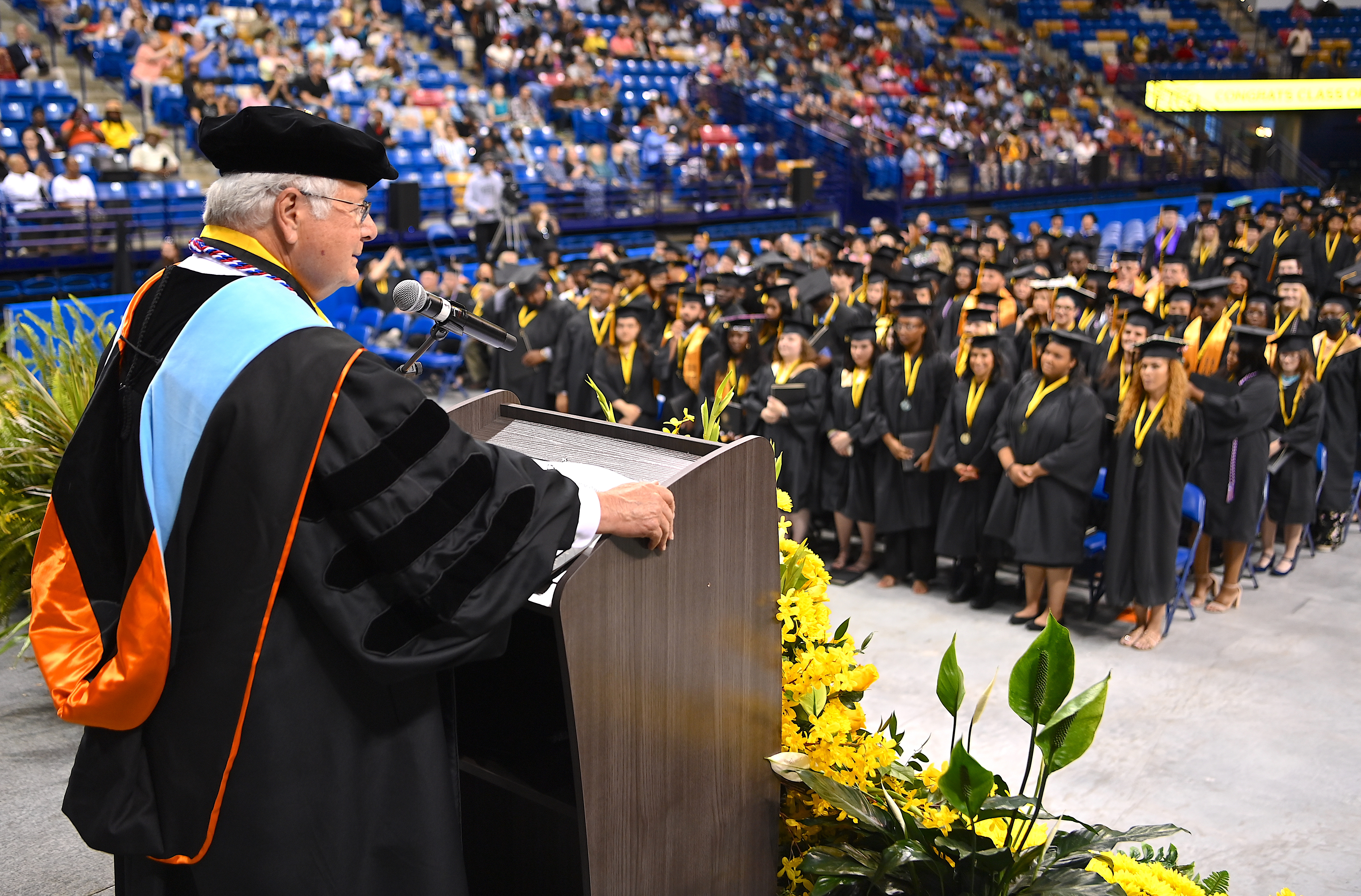 Dr. Larry Keen, photographed from behind, speaks at the podium. In the background, parts of the graduates and the audience are visible. 