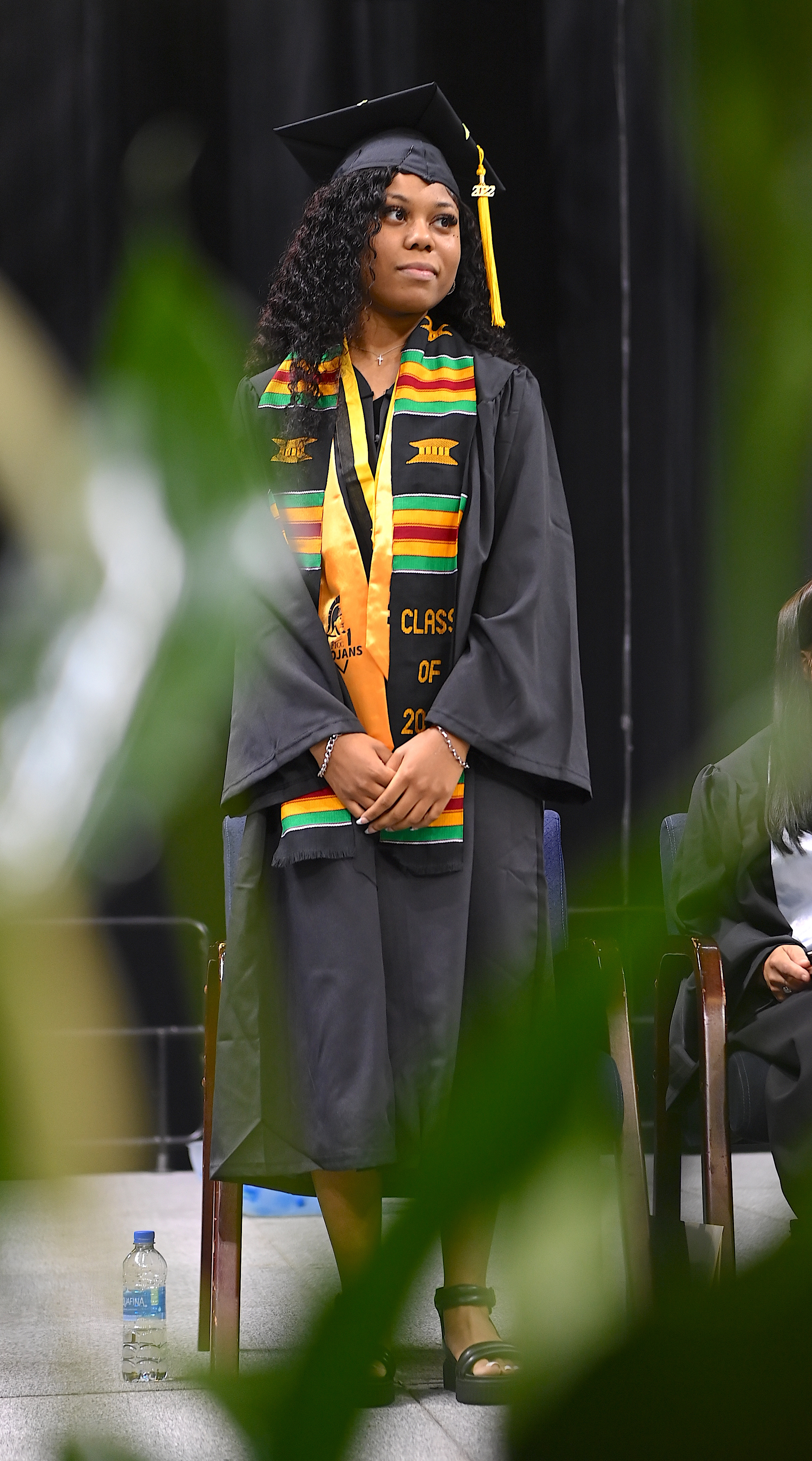 Kristen Woods stands on stage. She is wearing a both a gold stole and a black stole with green, yellow and red stripes. 