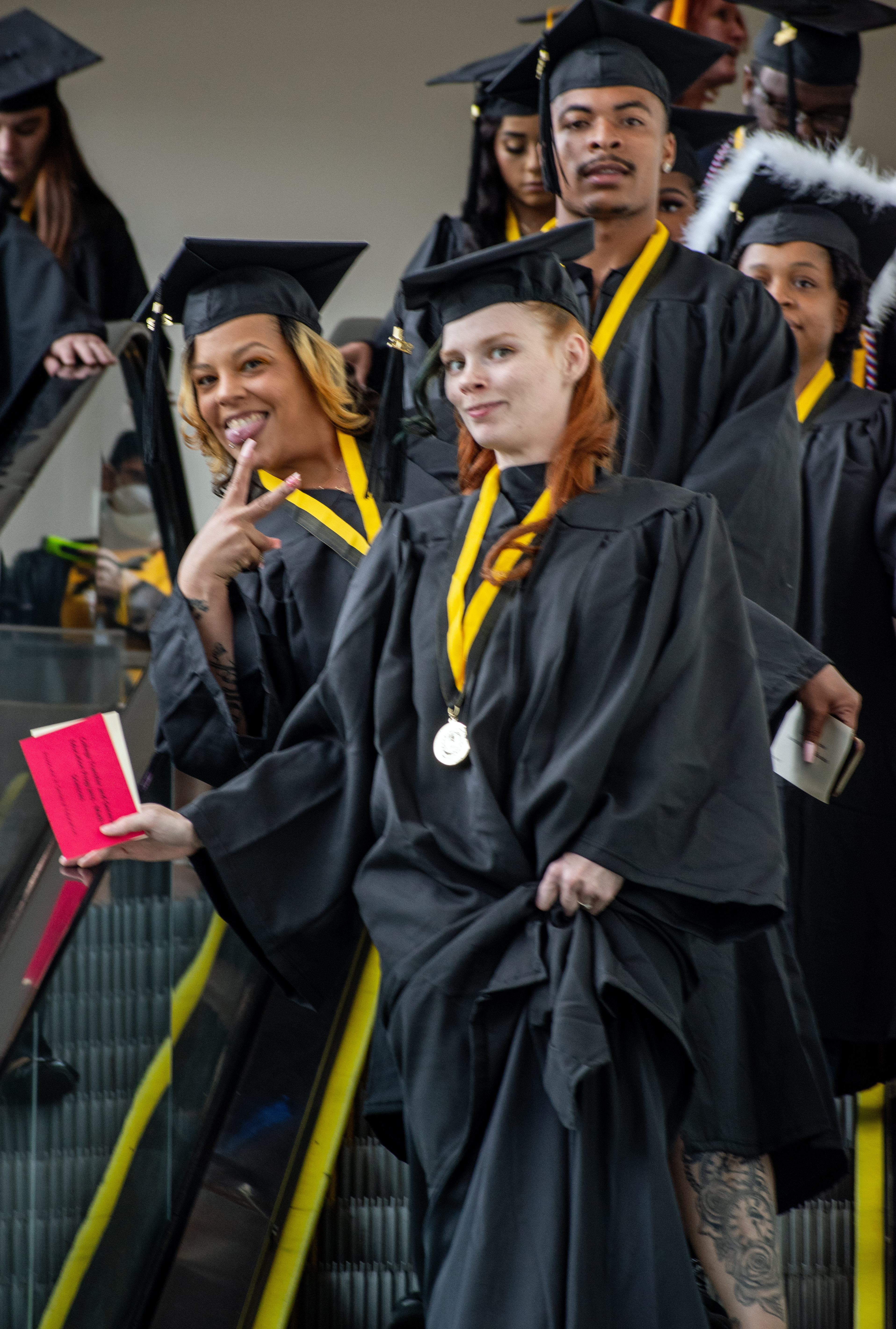 Two graduates at the front of the line descending the escalator smile for the camera. The second graduate is holding up two fingers in a peace sign.