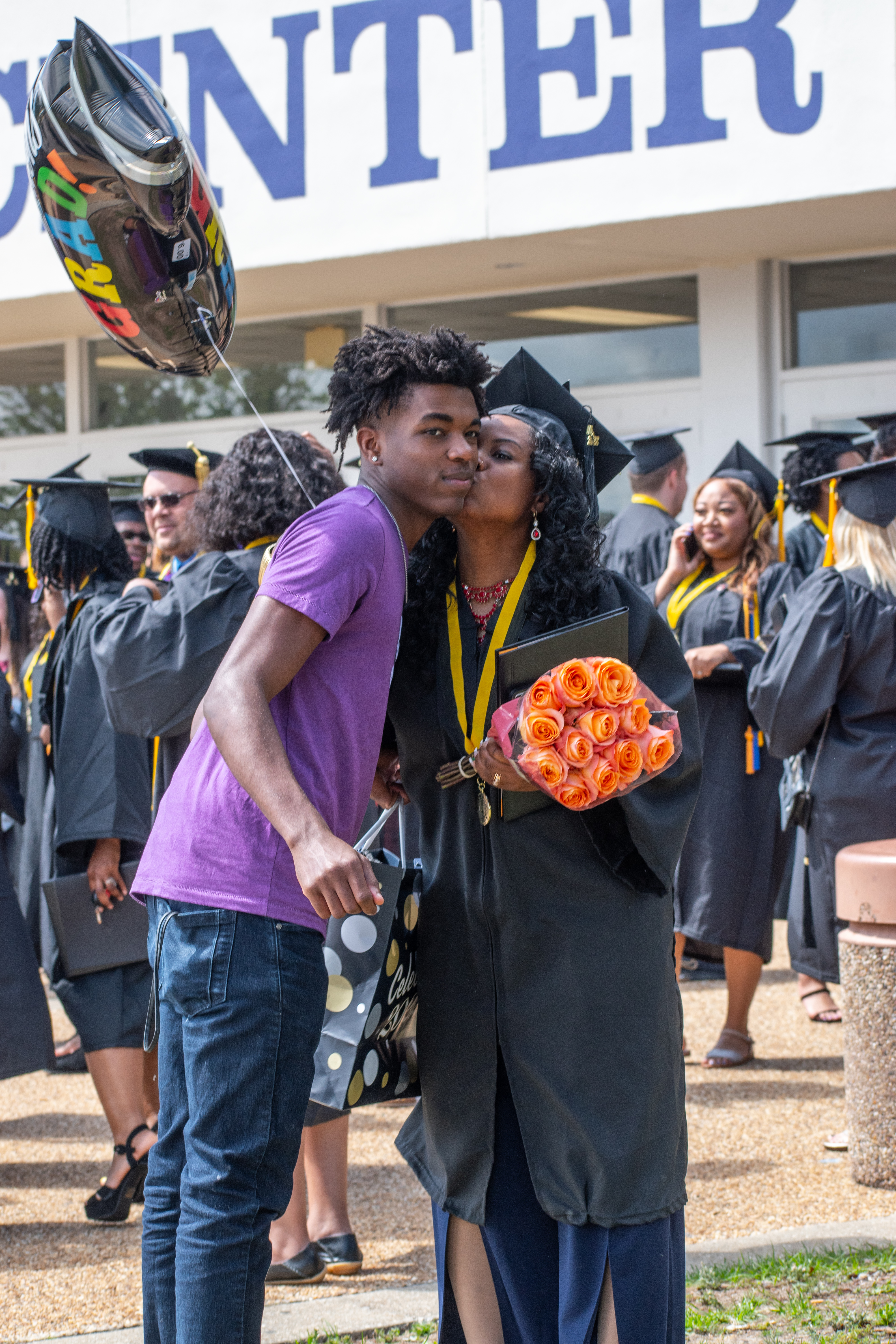 A graduate holding a bouquet of flowers kisses the cheek of a  young man. The pair are outside in a crowd of graduates.