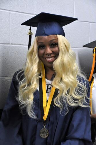 A close-up photo of a graduate with long blond hair. She is wearing dark blue cap and gown and a medal on a gold and black ribbon around her neck.