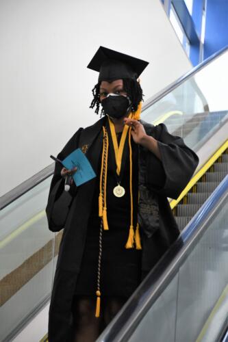 A graduate waves to the camera as she descends the escalator before graduation. She is wearing two gold cords, a black and gold cord and medal on a black and gold ribbon around her neck.
