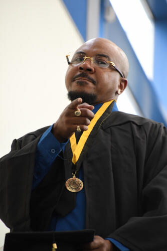 A graduate without his cap looks down into and points at the camera as he comes down the escalator.