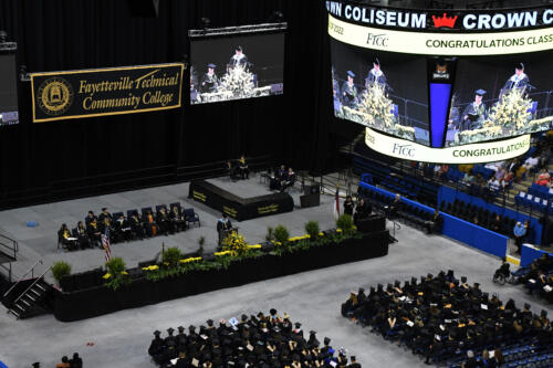 A wide photo looking down at the stage, the graduates seated in chairs on the arena floor and the jumbotron.