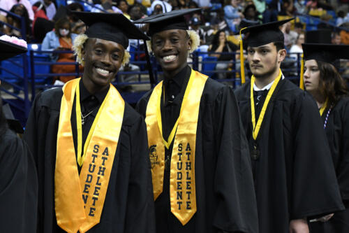 Twin brothers smile for the camera. They are wearing caps, gowns and gold stoles that say "Student Athlete." A third graduate stands slightly behind them.