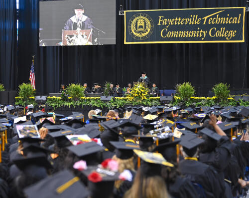 A wide photo of seated graduates, photographed from behind, look on as Dr. Keen speaks on stage during graduation.