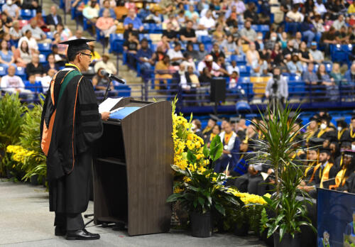 Dr. Choi, photographed from the side, speaks at the podium. Part of the graduates and the crowd is visible in the background.
