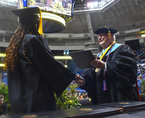 A graduate, photographed from behind, accepts her degree portfolio from Dr. Keen. 