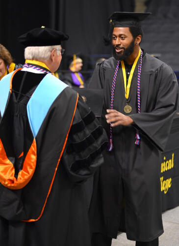 A tall graduate accepts his degree portfolio from Dr. Keen. He is wearing a red, white and blue cord and a medal on a black and gold ribbon around his neck. 