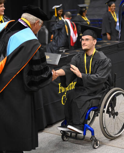 A graduate, utilizing a wheelchair, shakes hands with Dr. Keen and reaches for his degree portfolio.