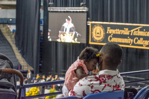 An audience member holds a young child in the foreground. In the background, part of the stage, the college banner and the projection screen are visible. 