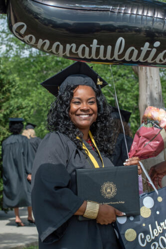 A graduate standing outside smiles at the camera while holding a bouquet of flowers.