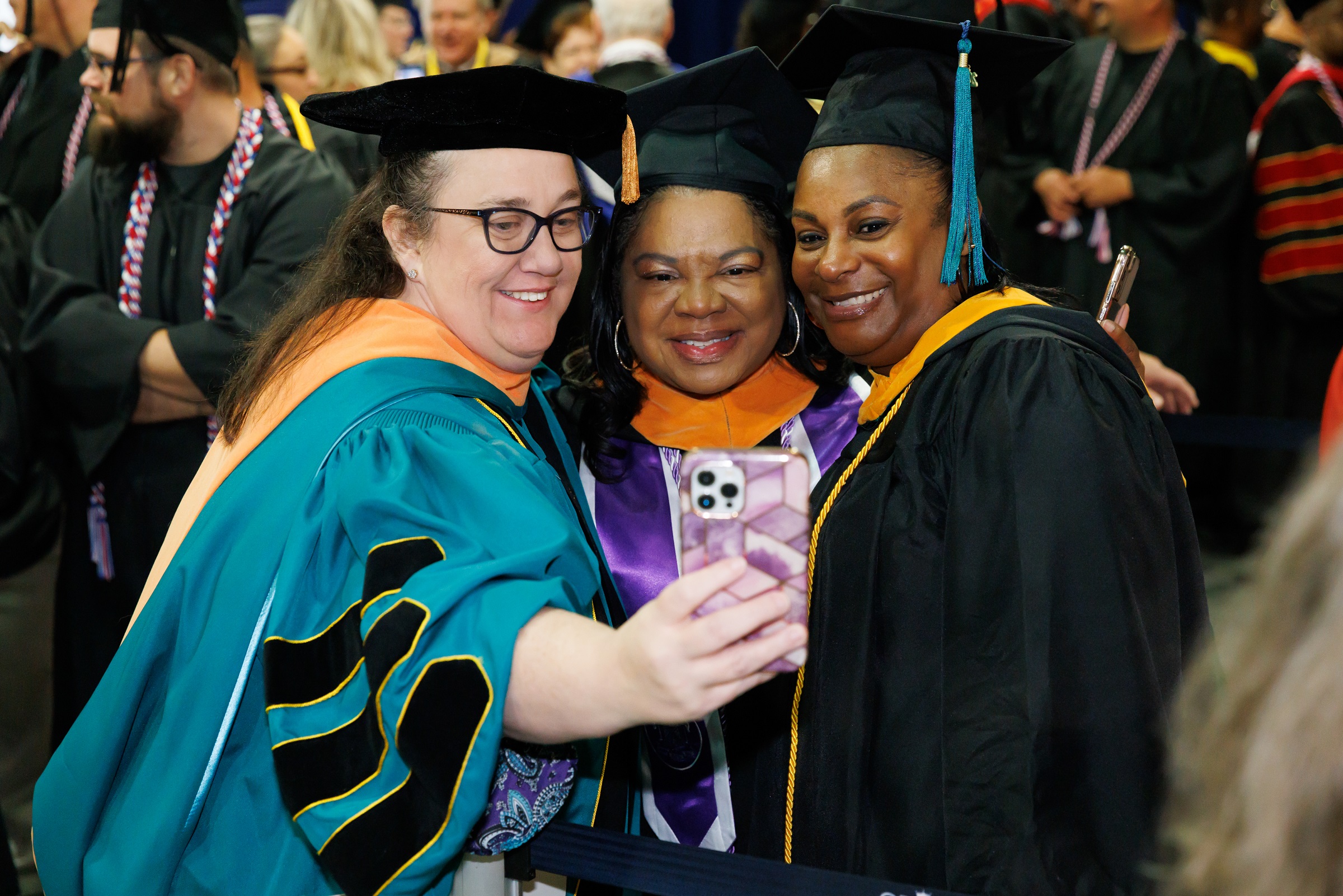 A faculty member takes a selfie with two other instructors. All of them are dressed in graduation regalia.