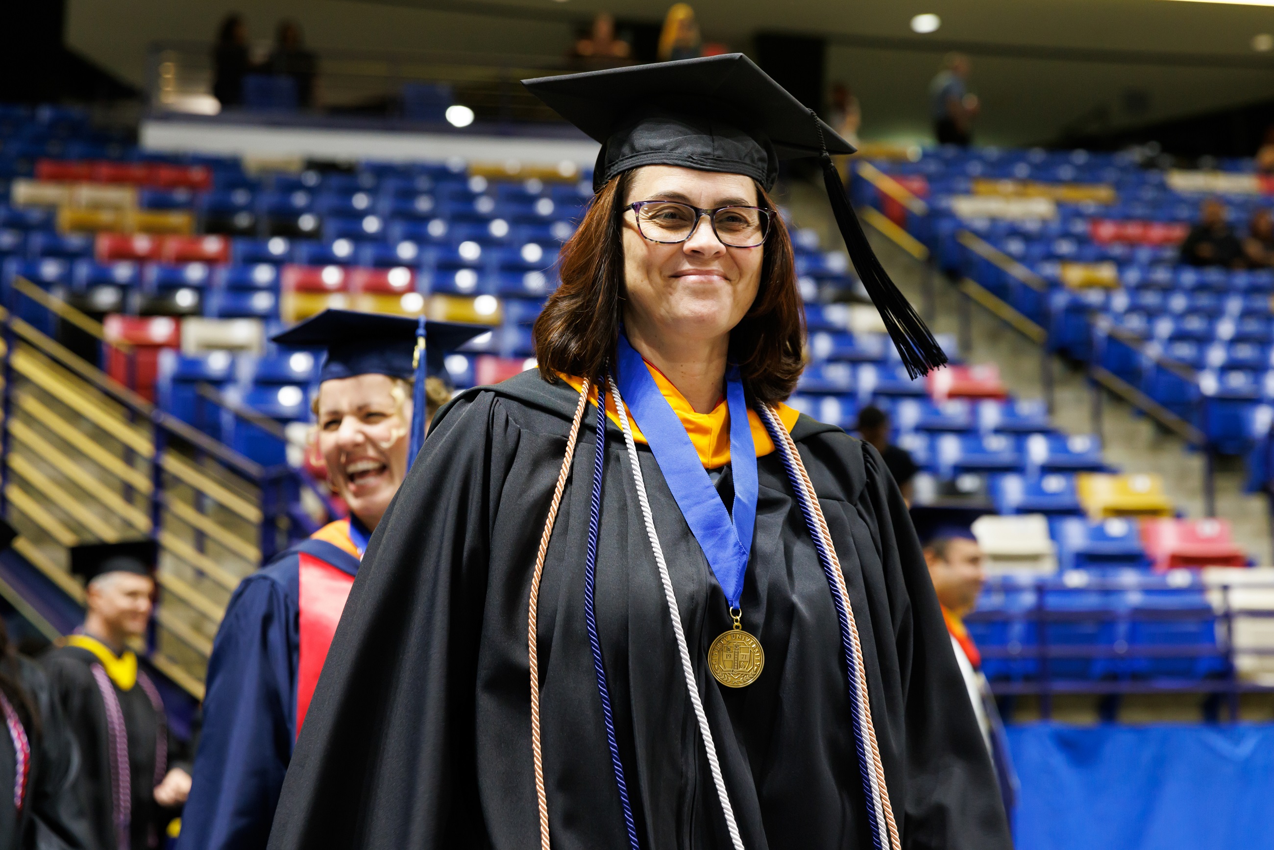 A faculty member dressed in graduation regalia smiles at the camera as she enters the Crown floor.