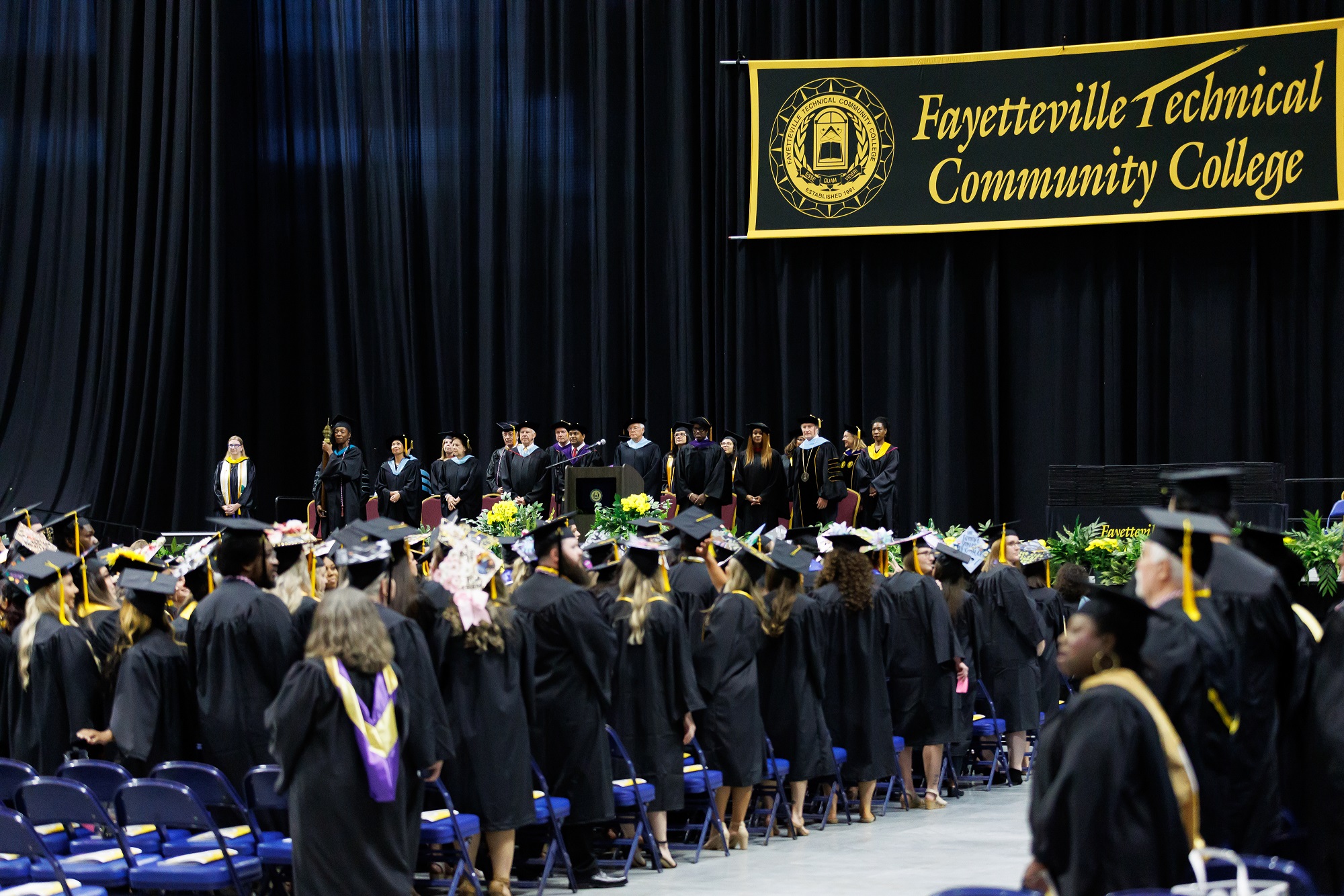 A wide photo taken from behind the standing group of graduates shows the distinguished guests standing on stage.