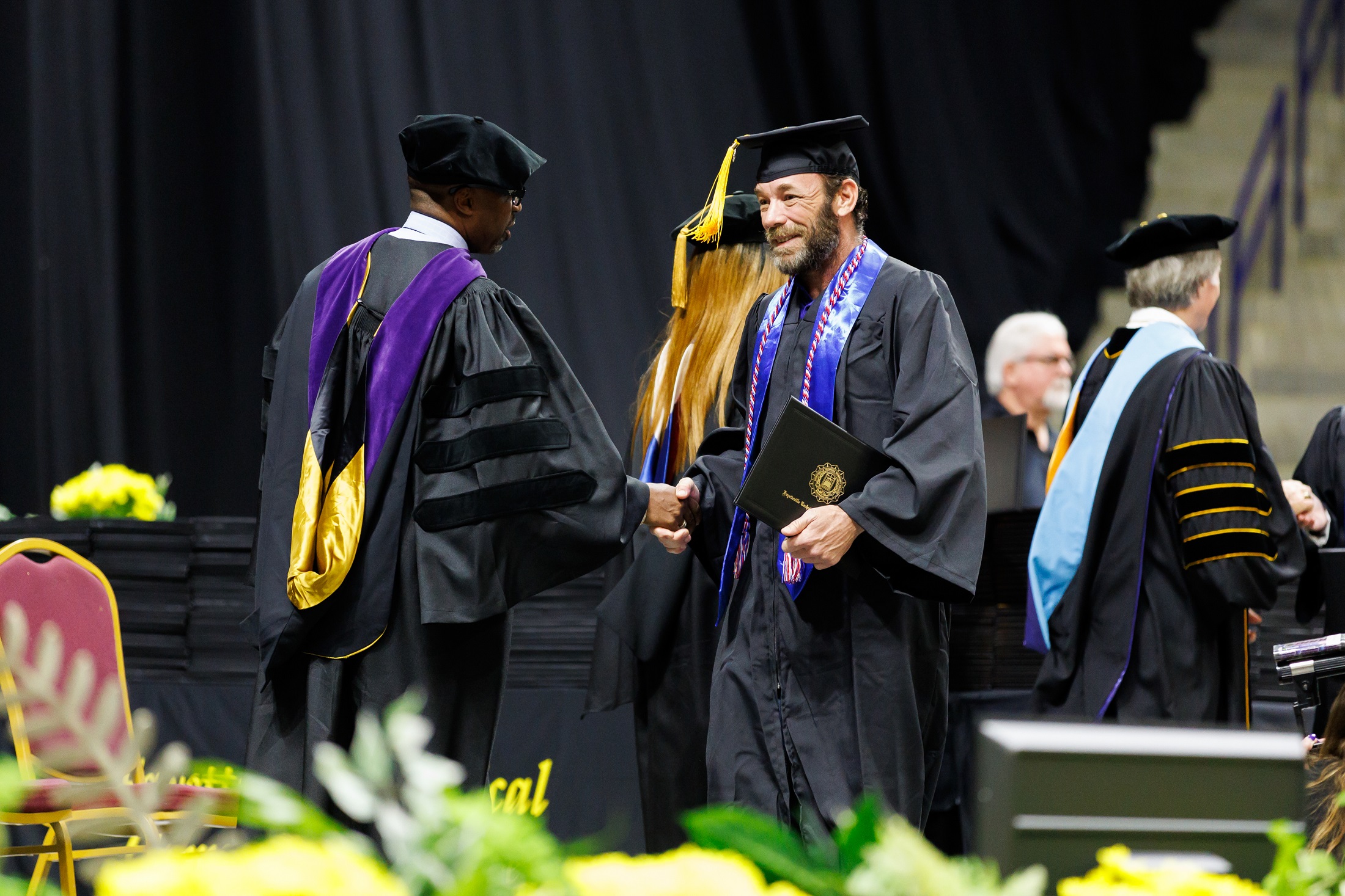 A graduate wearing a blue stole shakes hands with Ronald Crosby Jr. on stage.
