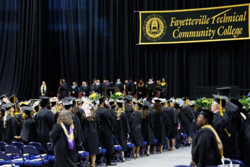 A wide photo taken from behind the standing group of graduates shows the distinguished guests standing on stage.