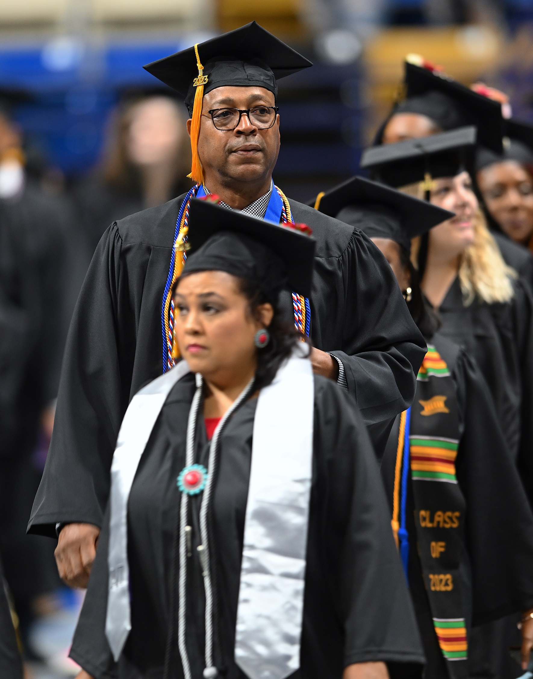 A graduate in glasses wearing multiple cords and a blue ribbon walks in the processional line.