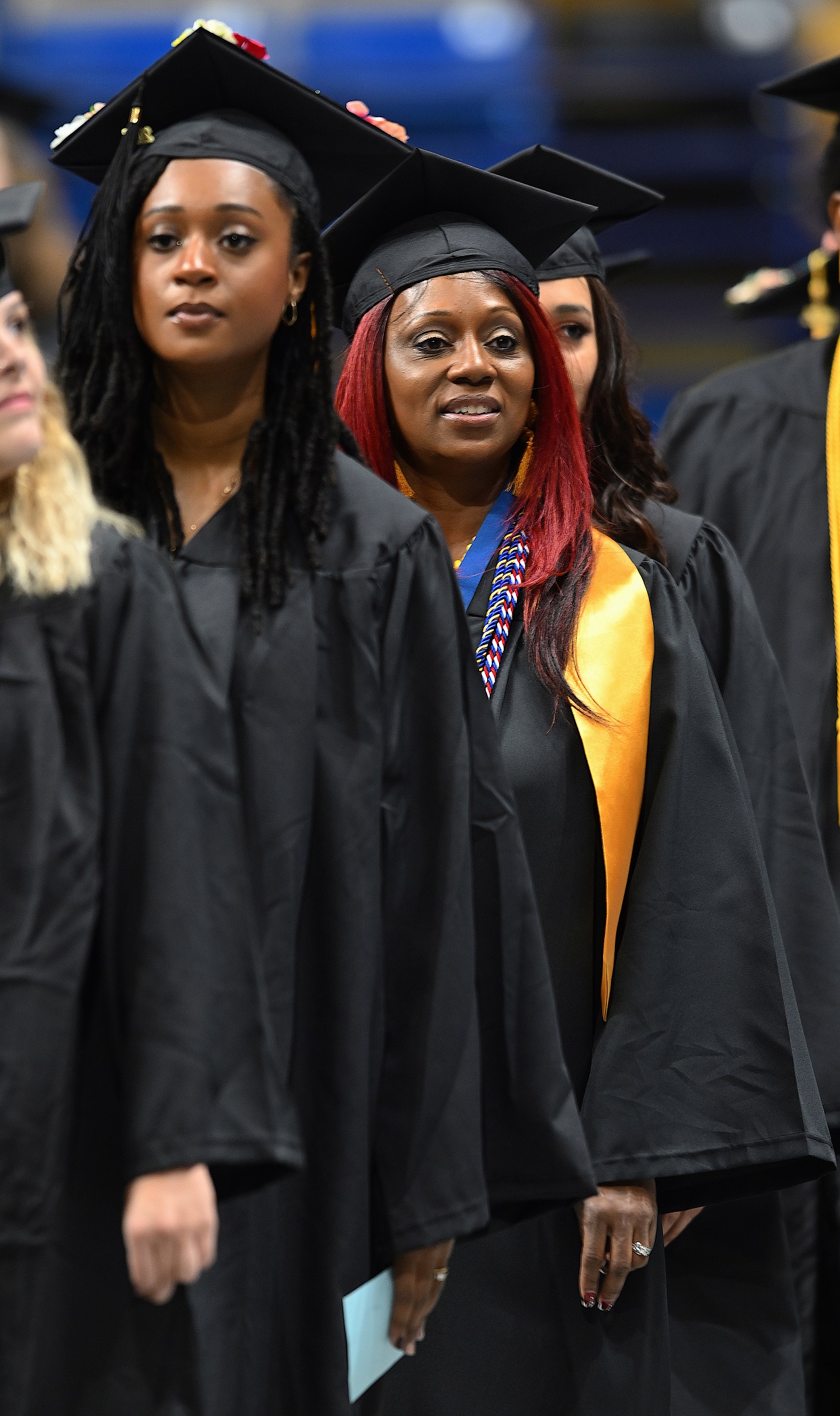 A graduate with red hair, wearing a gold stole, red, white and blue cord and a blue ribbon walks in the processional line.