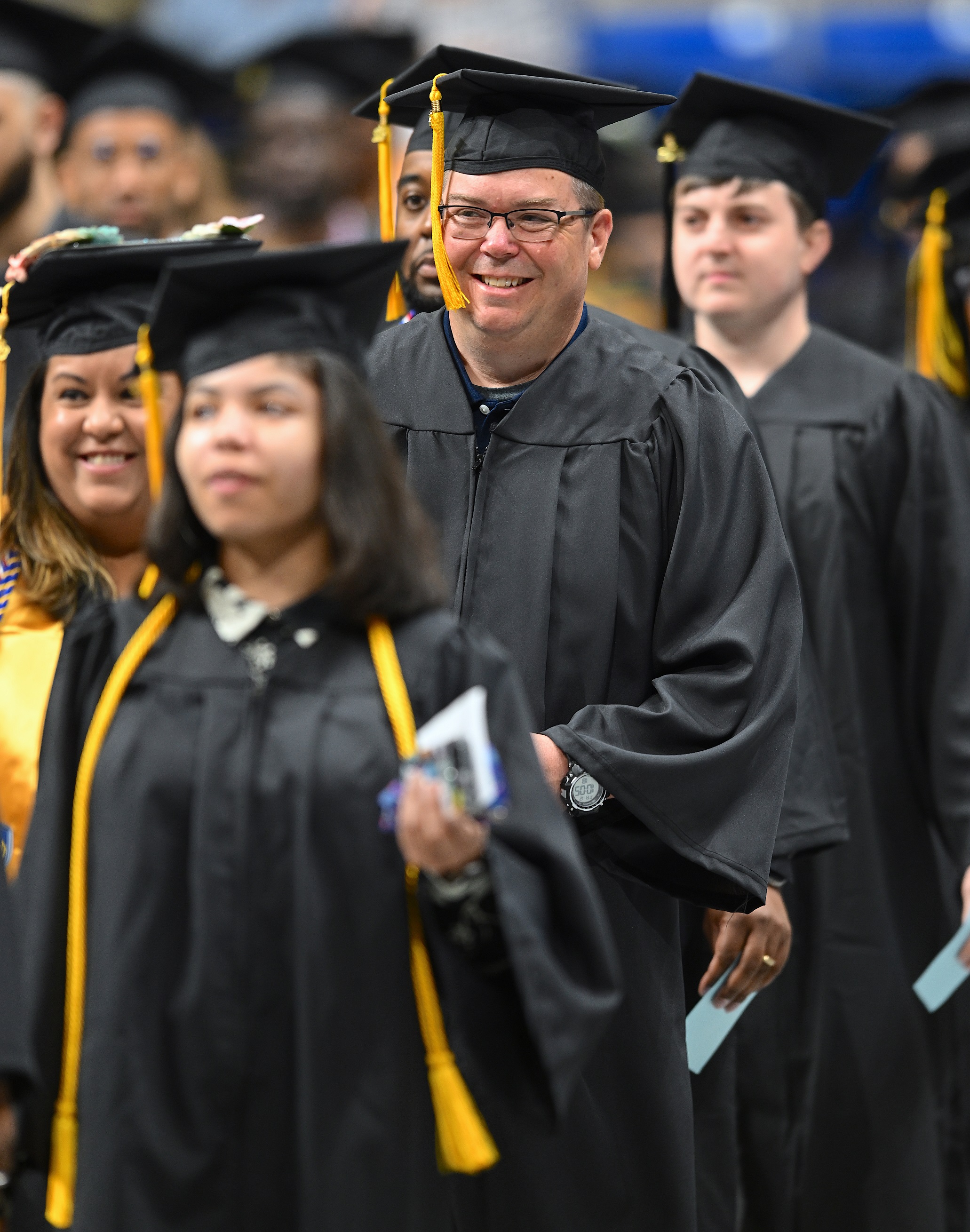 A graduate wearing glasses smiles as he walks in the processional line.