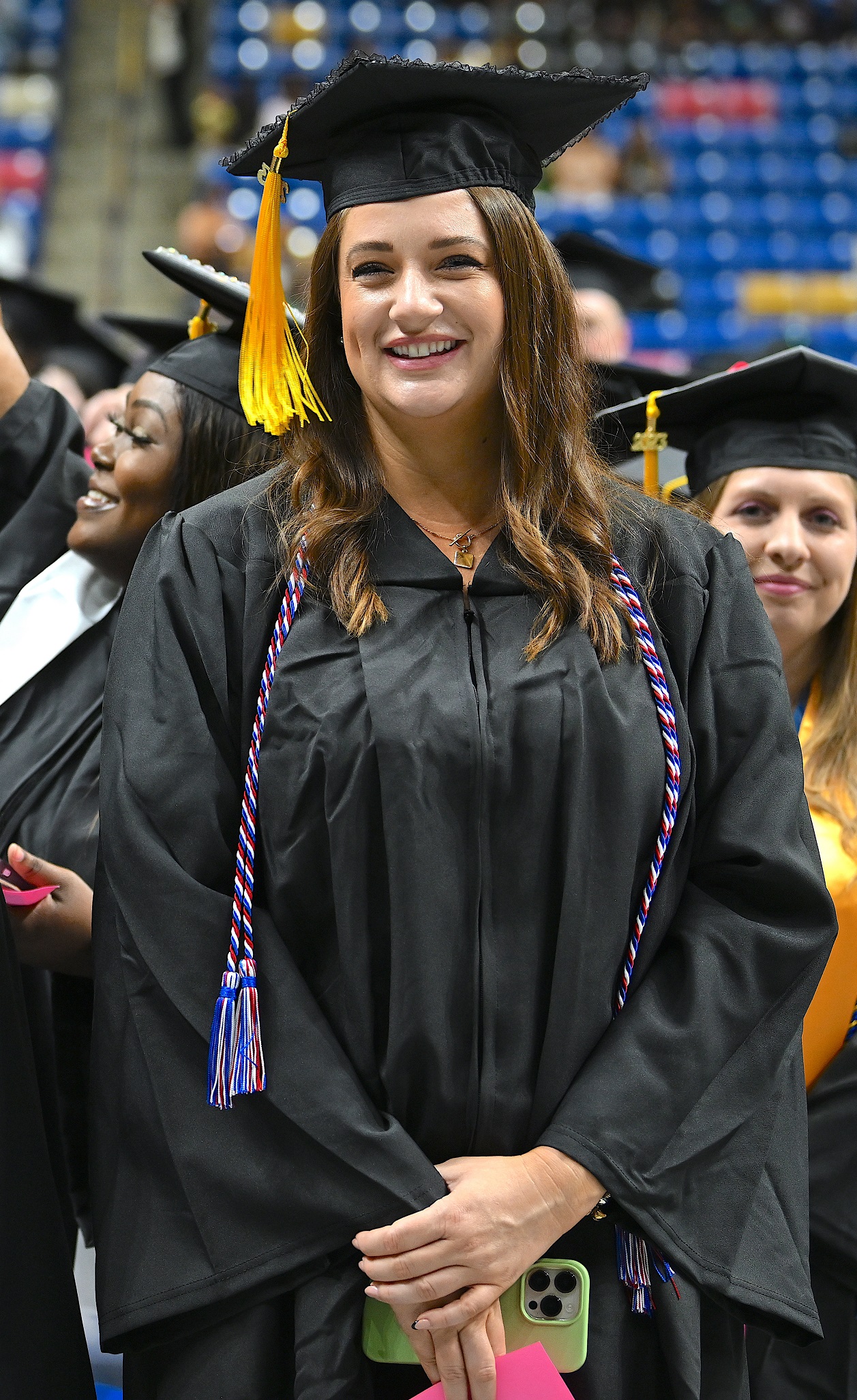 A graduate wearing a red, white and blue cord smiles at the camera while she waits to be seated.