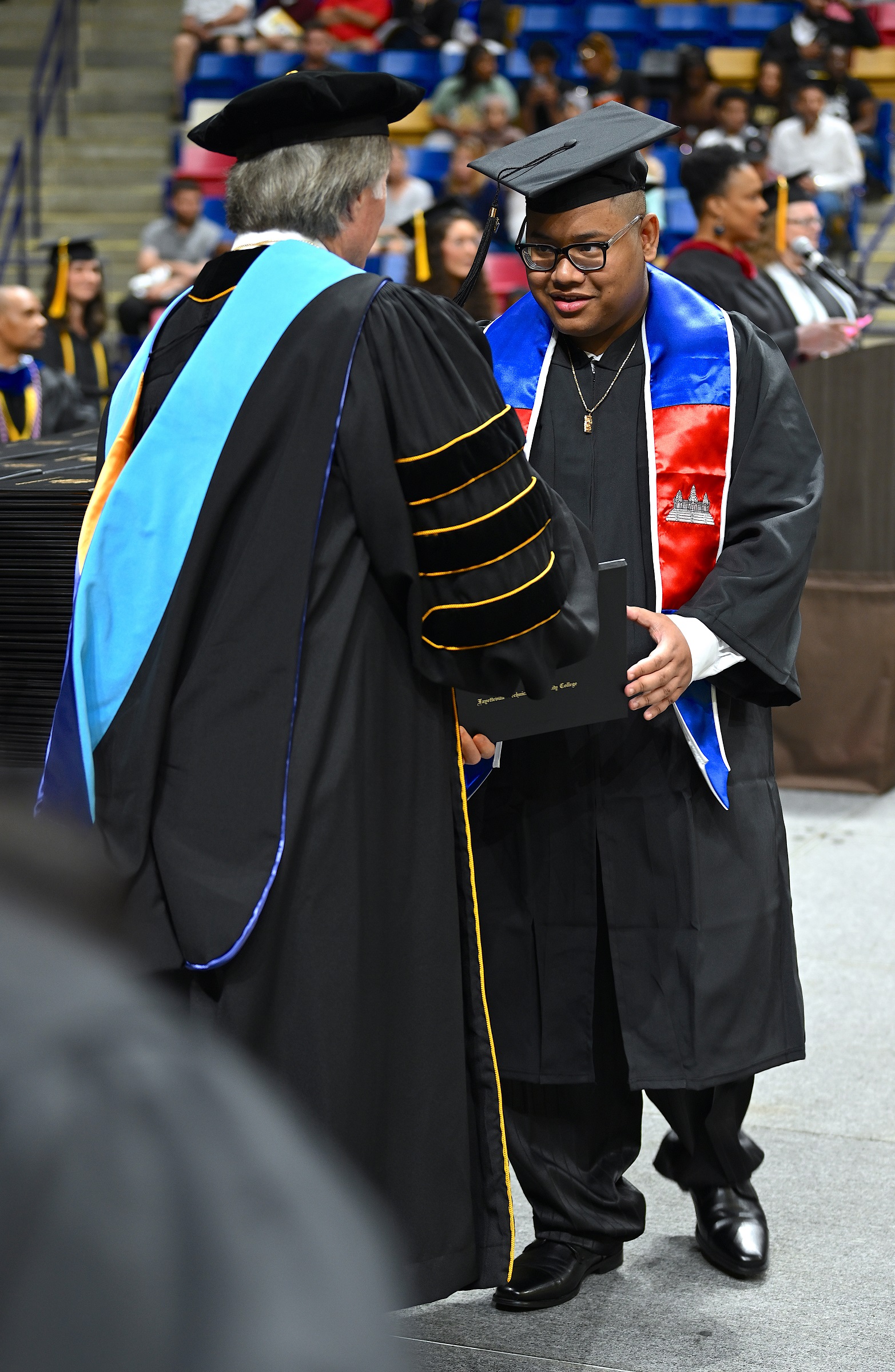 A graduate wearing a stole depicting the Cambodian flag accepts his diploma folder from Dr. Mark Sorrells.