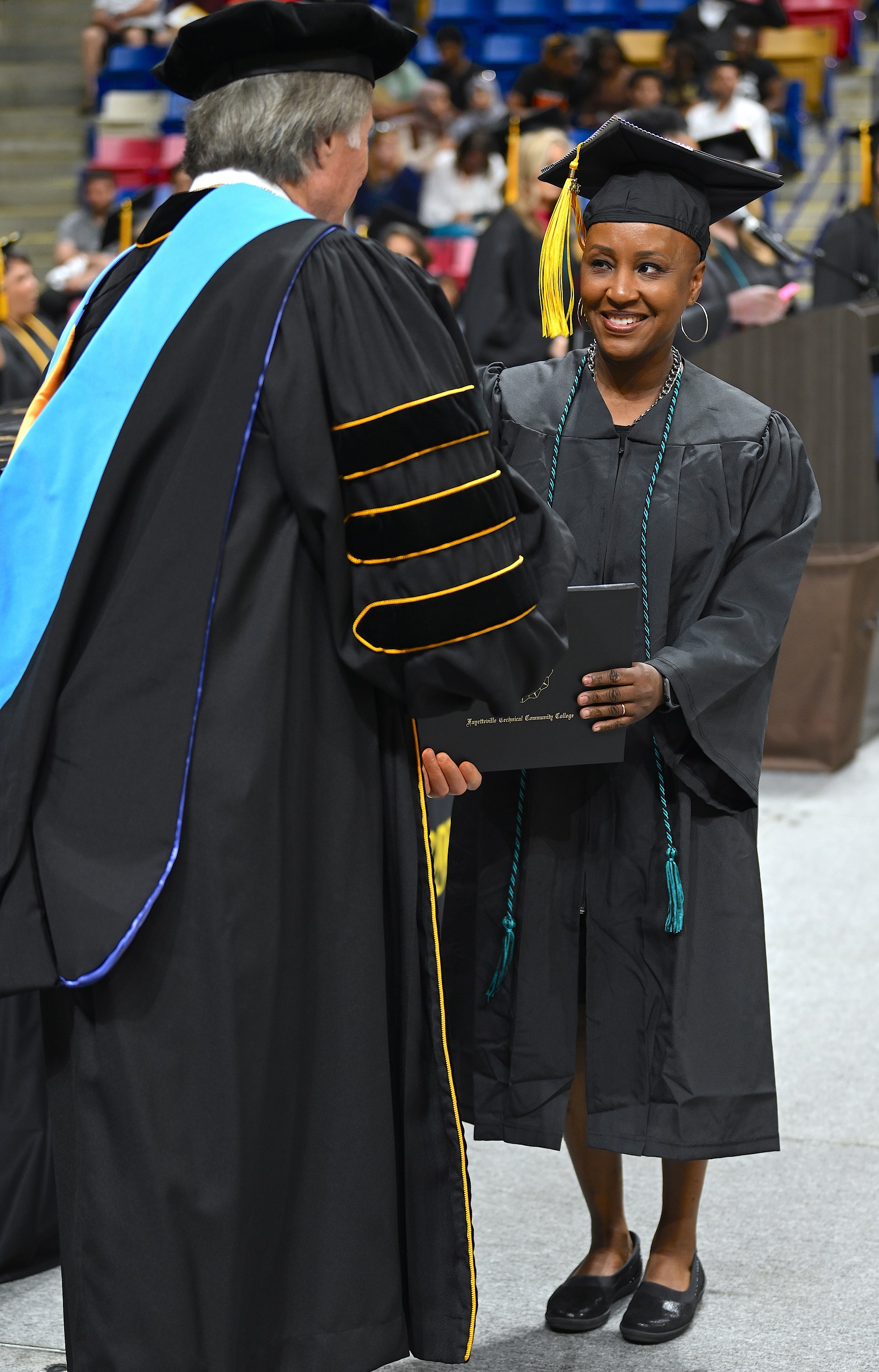 A graduate wearing a teal and black cord smiles at Dr. Mark Sorrells as she accepts her diploma folder from him.