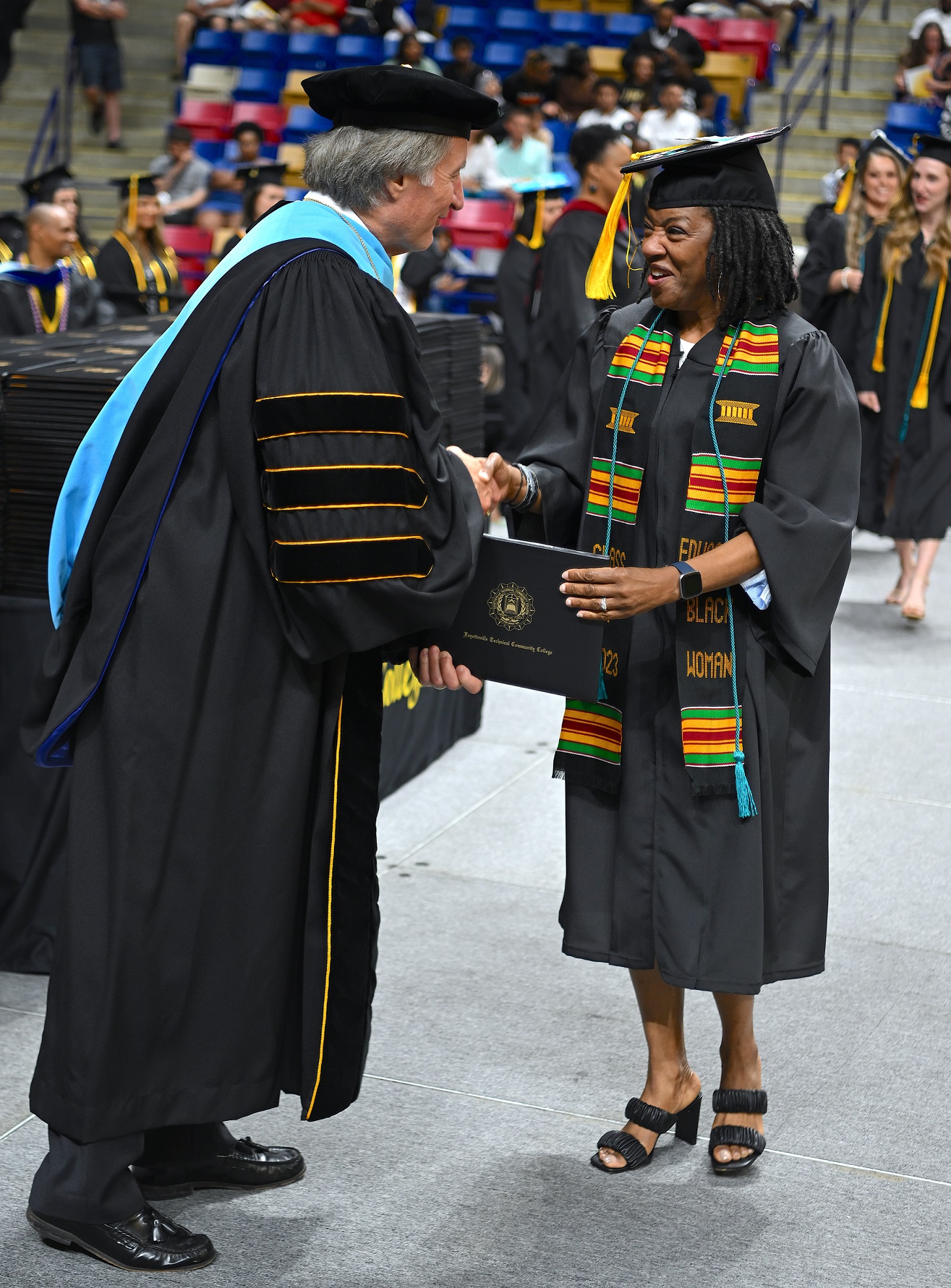 A graduate wearing a black, green, red and yellow stole smiles at Dr. Mark Sorrells as she accepts her diploma folder from him.