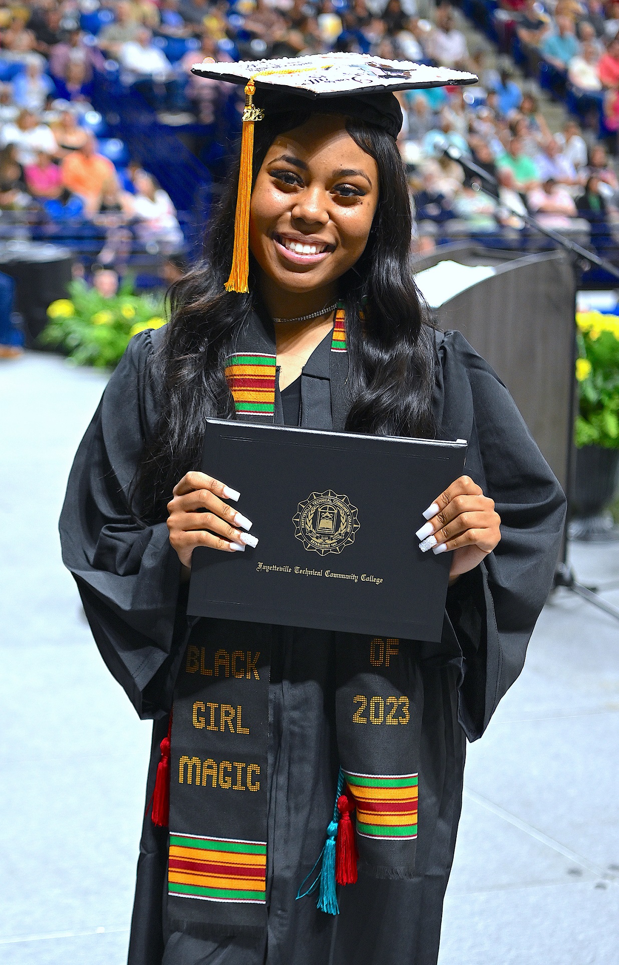 A graduate wearing a black, green, red and yellow stole smiles holds up her diploma folder as she smiles for the camera.