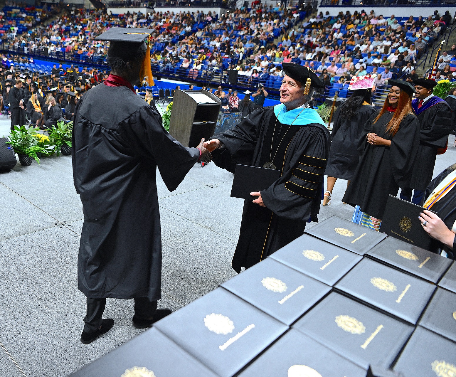 A photo taken from behind the table of diploma folders shows a graduating shaking hands with Dr. Mark Sorrells.