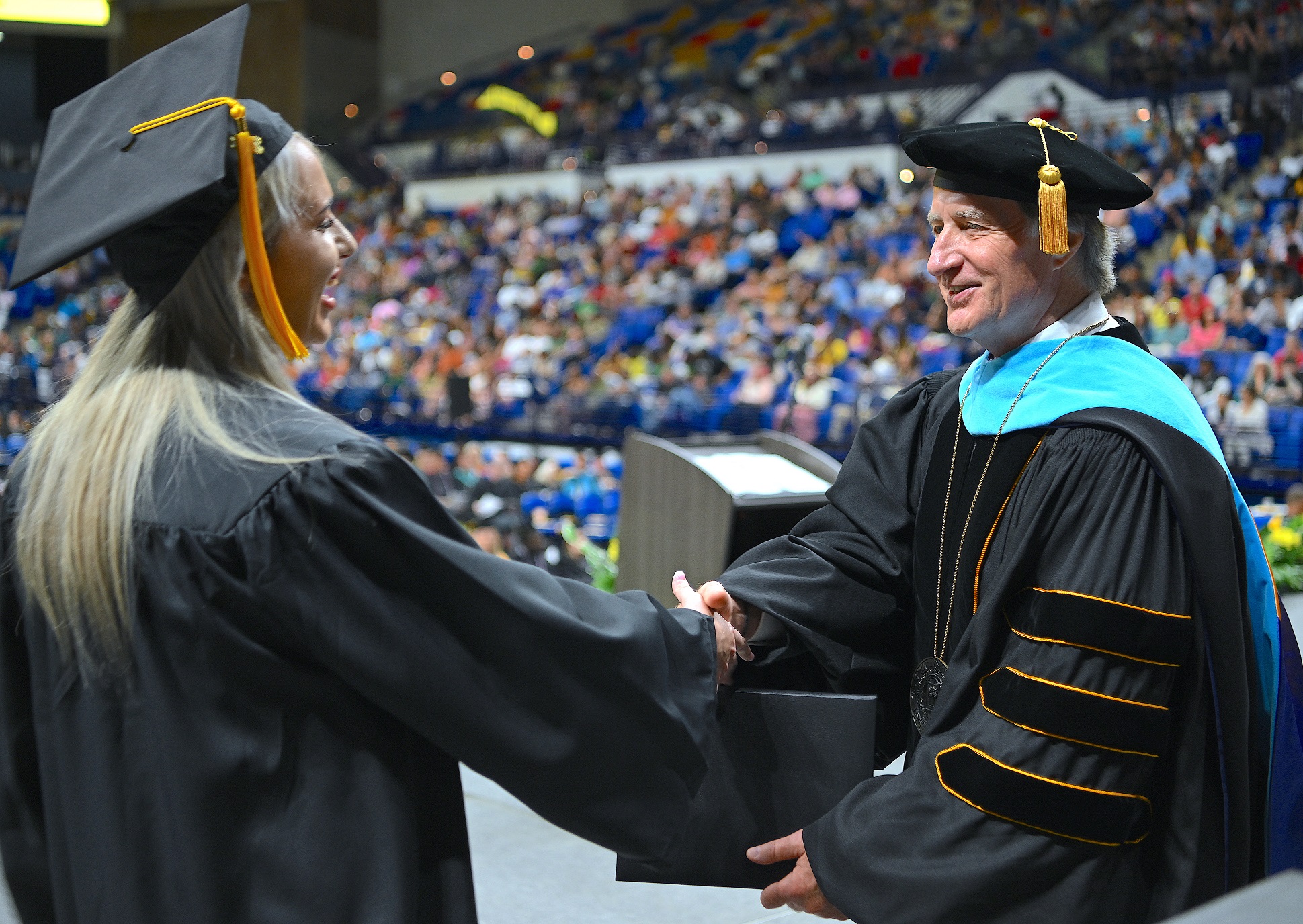 A graduate with long hair shakes hands with Dr. Mark Sorrells as she accepts her diploma folder from him.