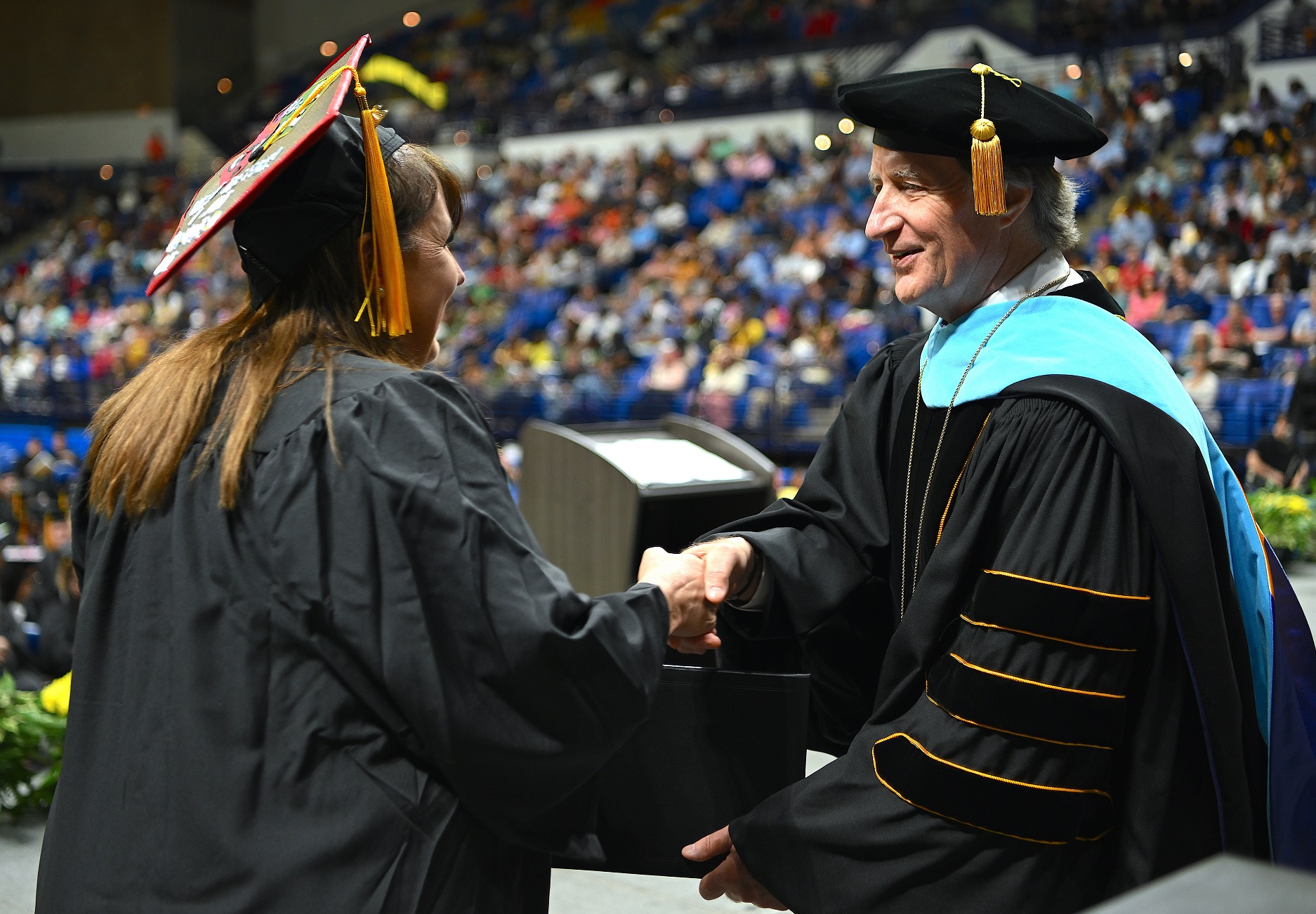 A graduate with long hair and a decorated cap shakes hands with Dr. Mark Sorrells as she accepts her diploma folder from him.