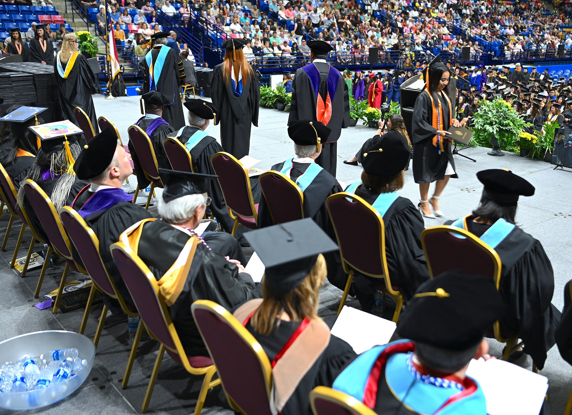A photo taken from behind the distinguished guests on stage, showing a graduate exit the stage after receiving her diploma folder.