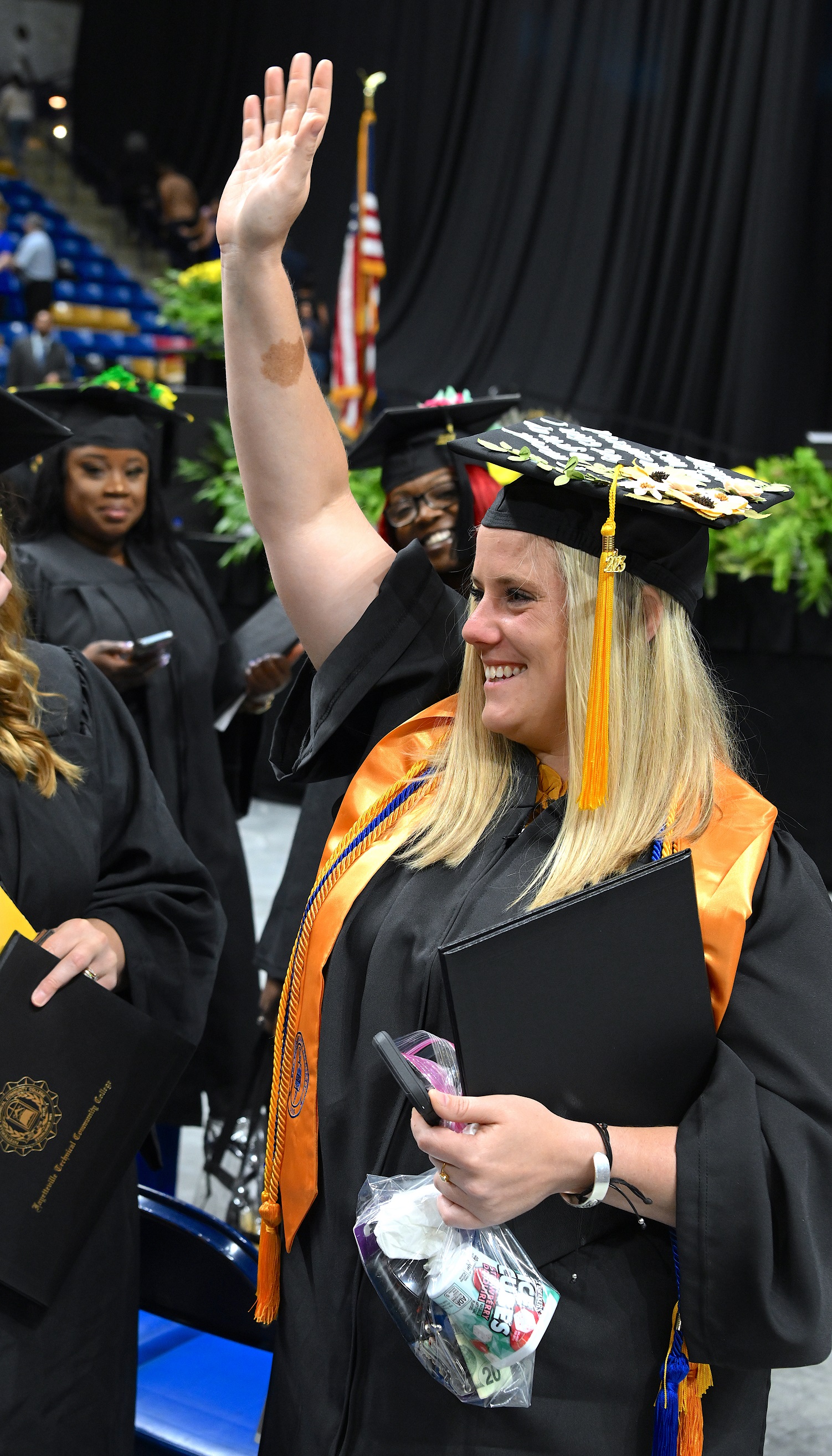 A graduate wearing a gold stole raises her hand to wave at someone in the crowd.