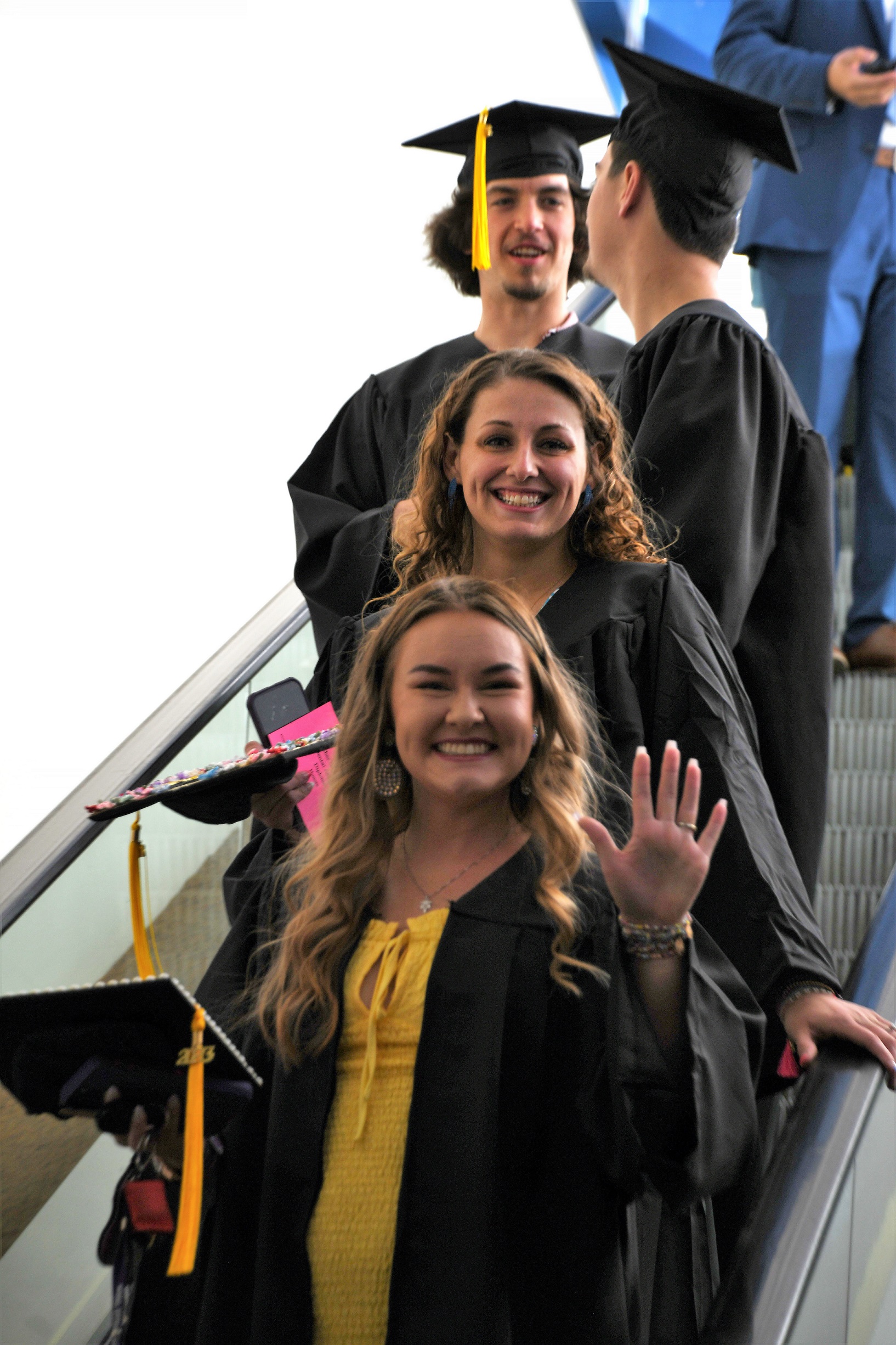 Graduates smile and wave as they descend the escalator on the way to line up for graduation.