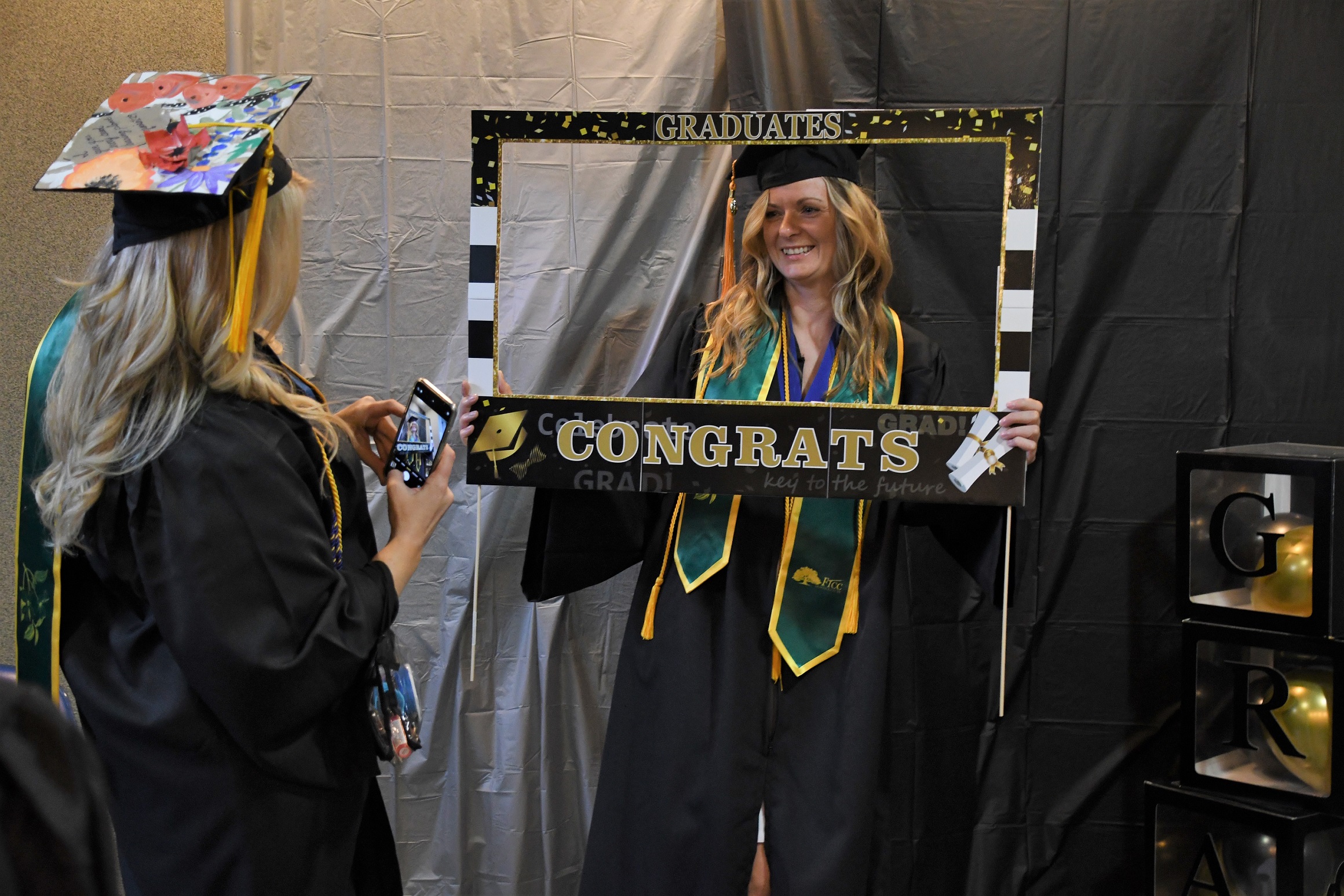 A graduate poses inside a frame that says "Congrats" as a classmate prepares to take a photo.