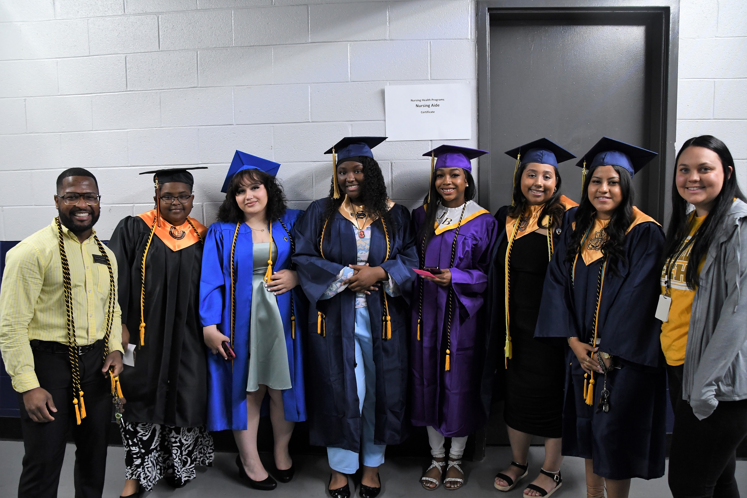 A group of graduate in different colored caps and gowns pose for a photo with a staff member on each side of the group.