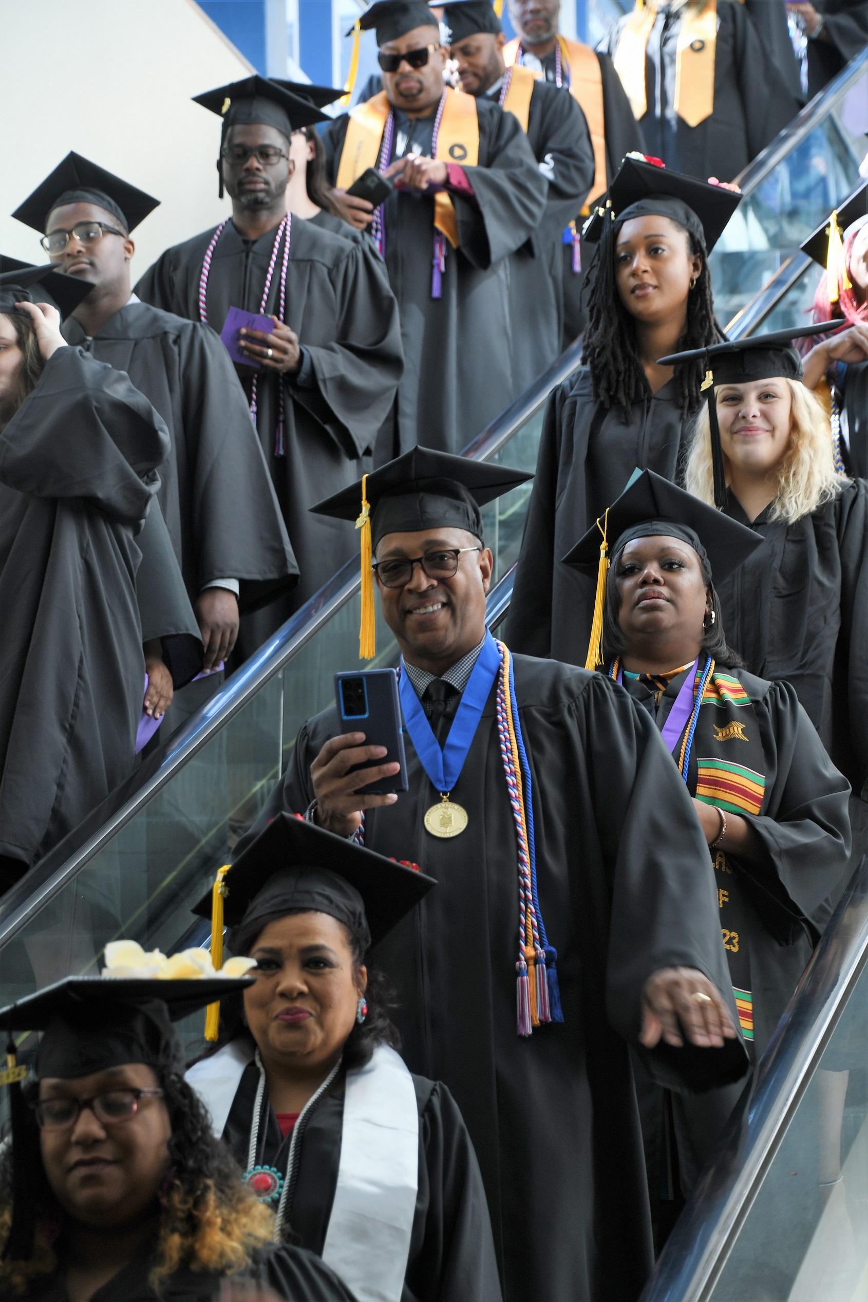 A graduate holds up his phone as he descends the escalator.