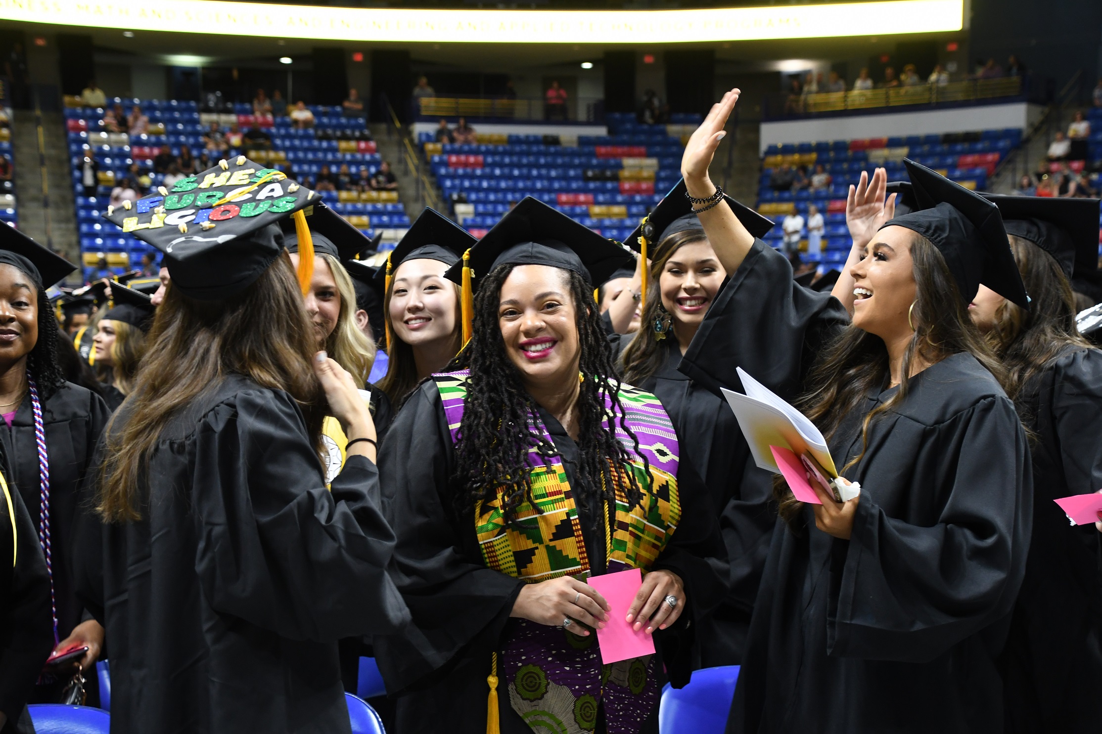A graduate in a group raises her hand to wave to someone in the crowd.