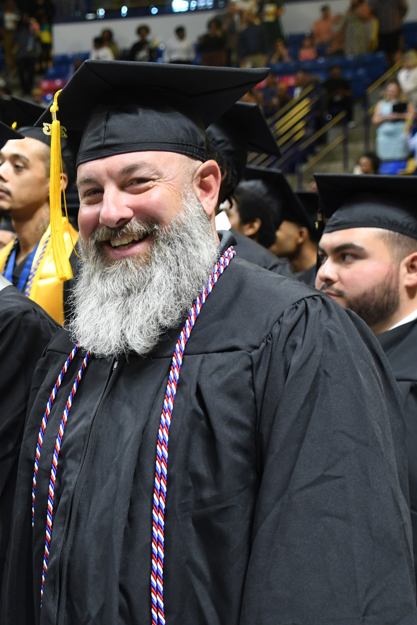 A graduate with a gray beard wearing a red, white and blue cord smiles at the camera while he waits to be seated.