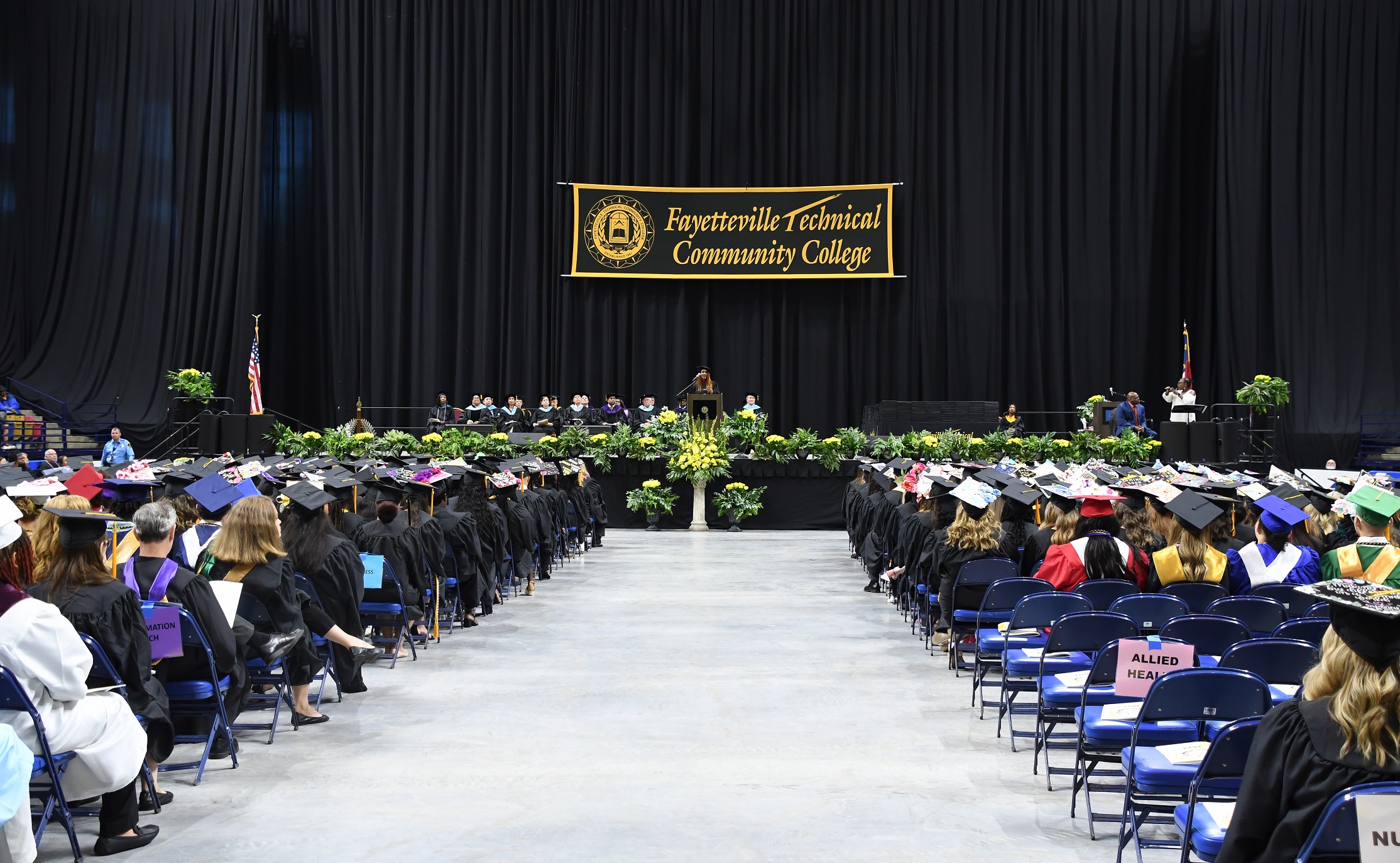 A photo taken from behind the center aisle that shows rows of graduates sitting on either side and stage in the background.
