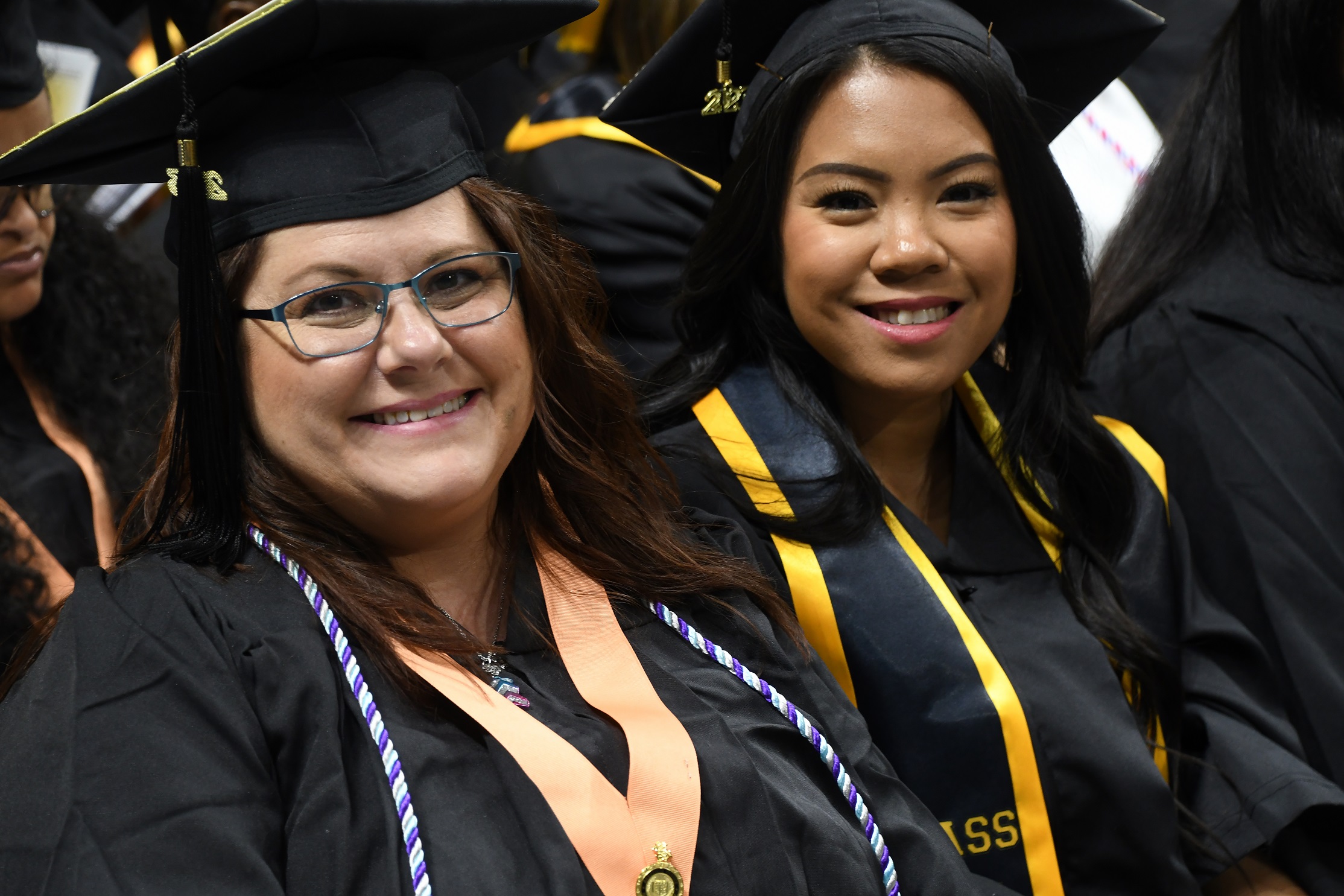 Two graduates, one wearing a peach-colored ribbon with a pin and one wearing a black and gold stole, smile at the camera.