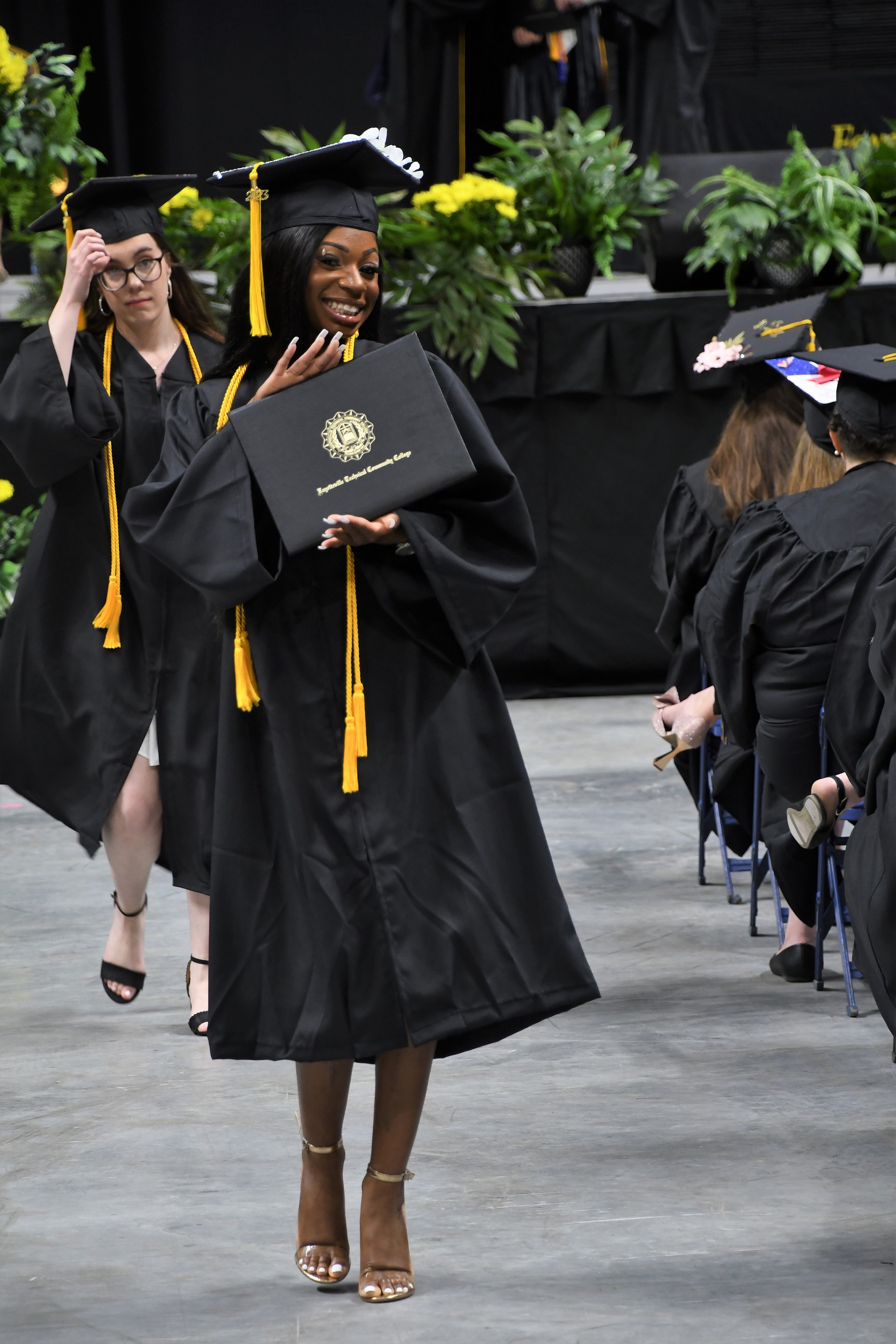 A graduate smiles and displays her diploma folder as she walks back to her seat.
