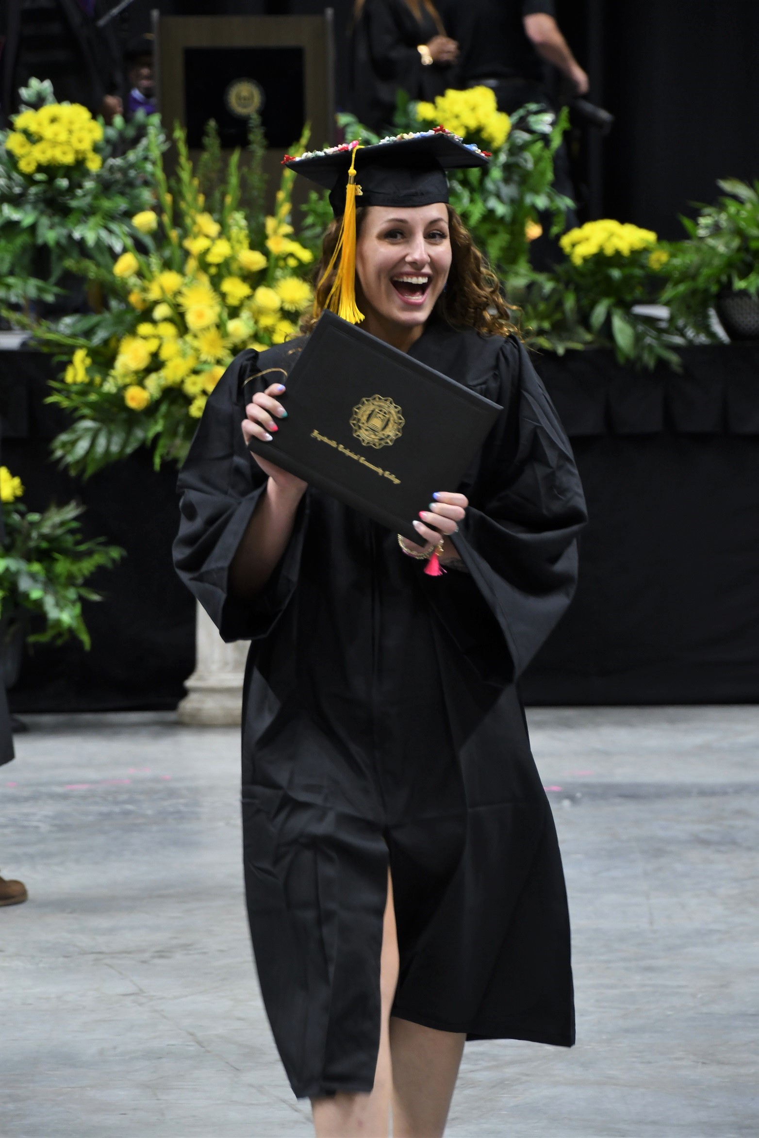 A graduate smiles and holds up her diploma folder as she walks back to her seat.