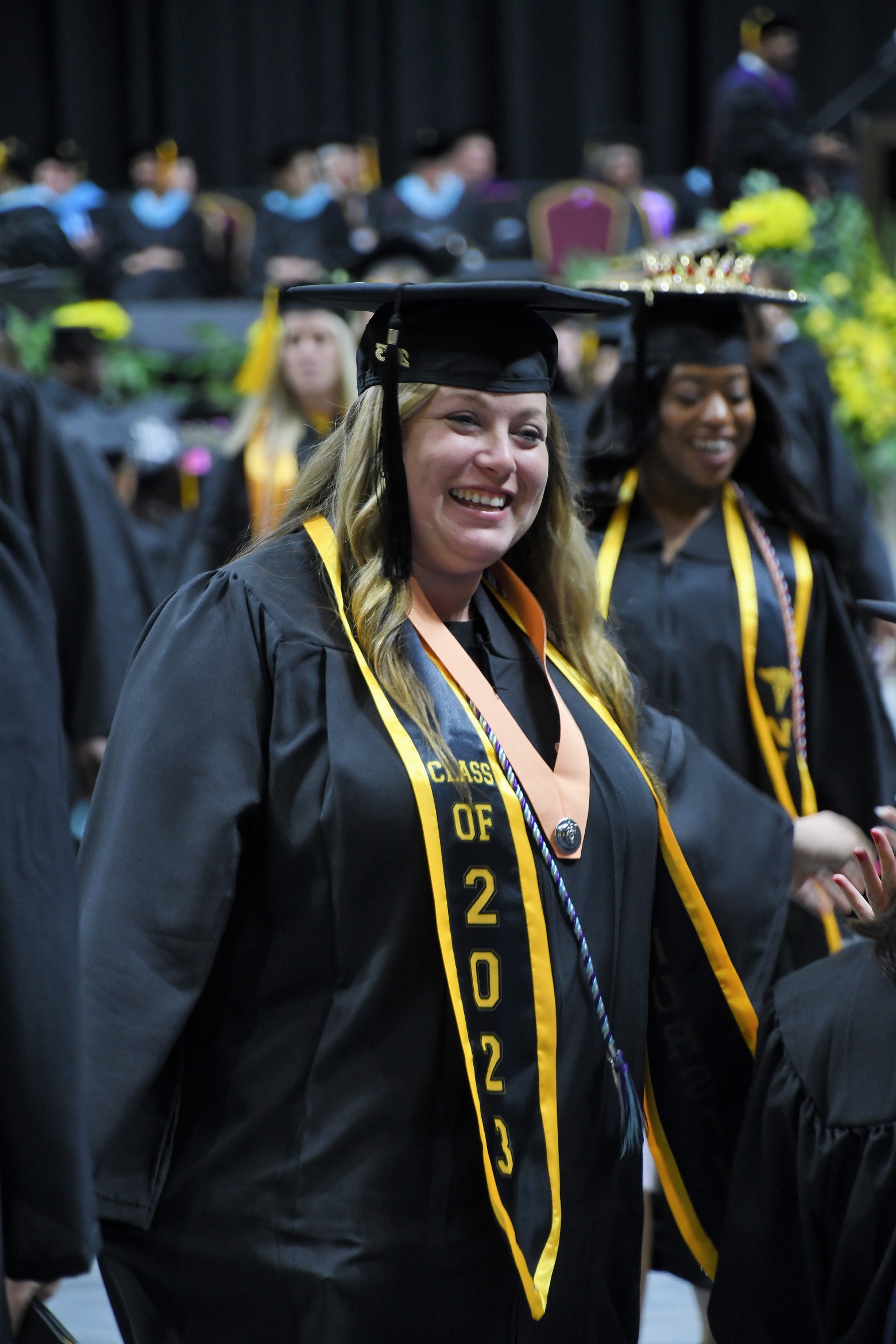 A graduate wearing a black and gold stole and a peach-colored ribbon with a pin smiles.